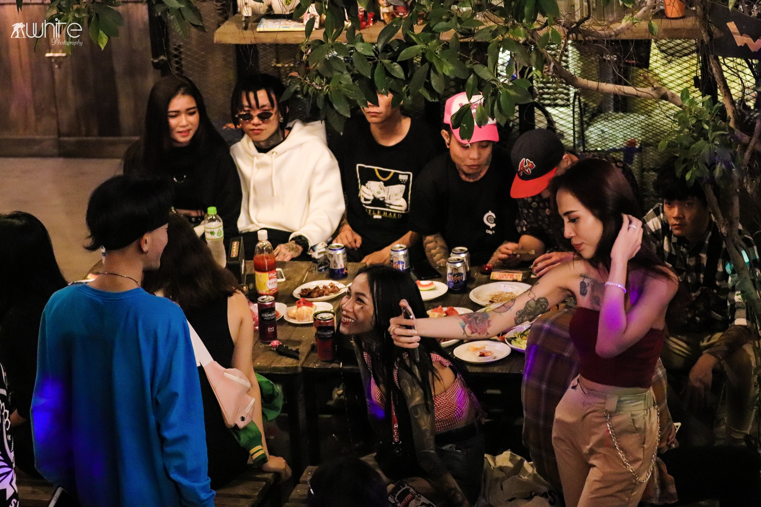 Group of people sitting around a table enjoying food and drinks at a social gathering in a dimly lit indoor setting with greenery.