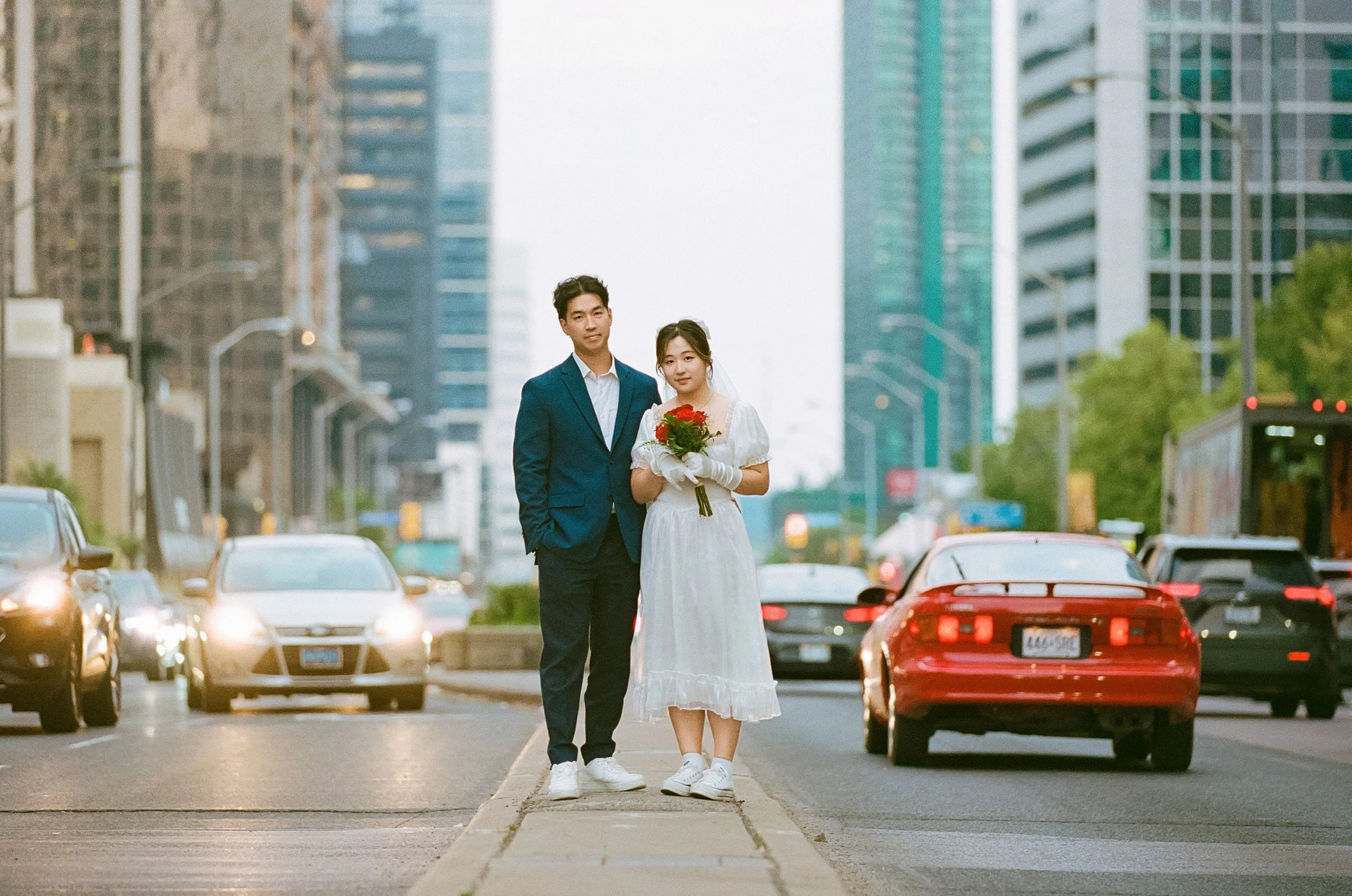 A young couple dressed for a wedding stands on a city street with high-rise buildings and cars around them. The bride holds a bouquet of red flowers, and the groom wears a blue suit.