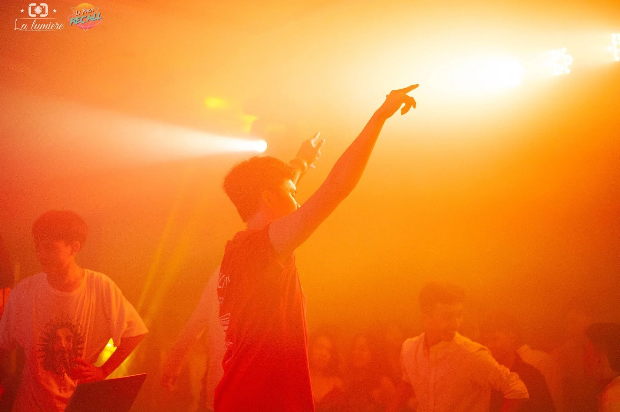 A young person with curly hair dancing or performing at a party or concert with orange and yellow lighting, and other people in the background.