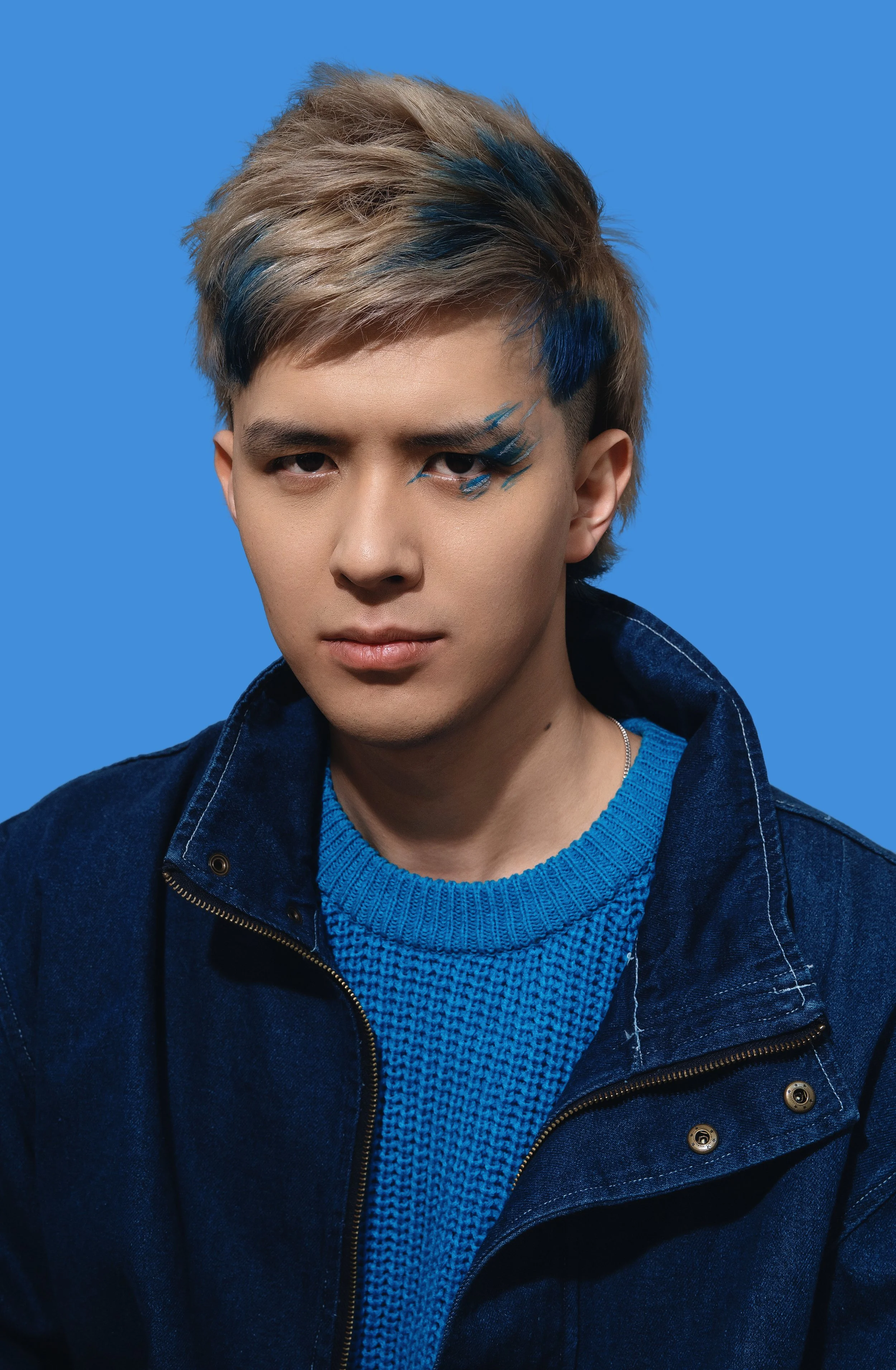 Portrait of a young man with styled hair featuring blonde and blue streaks, wearing a blue knit sweater and denim jacket, with a blue background.