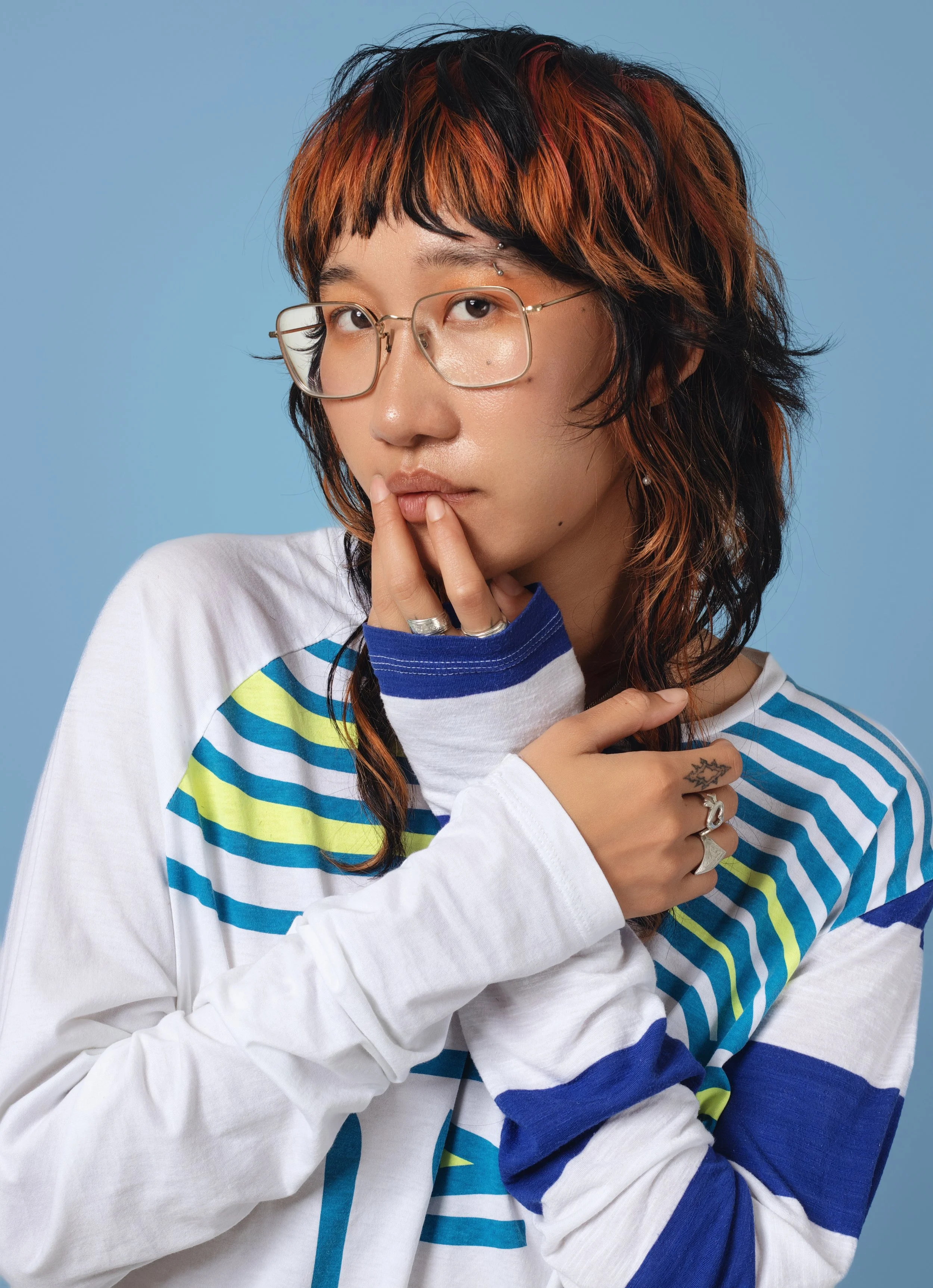A young woman with short, wavy, reddish-black hair and glasses, wearing a colorful striped shirt, posing against a plain blue background, with her hand near her face touching her lips and chest.