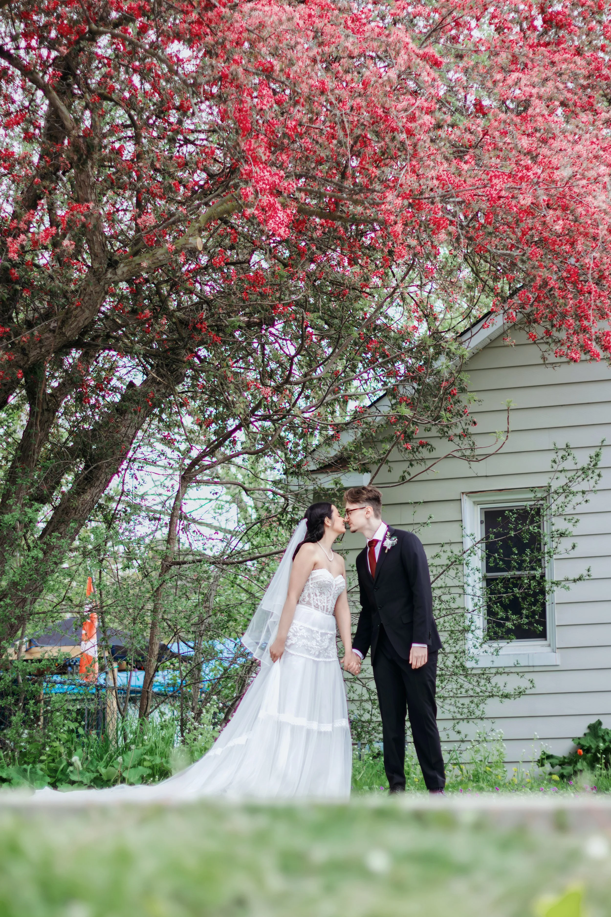 A bride and groom in wedding attire holding hands under a flowering tree, near a house with white siding and a window.