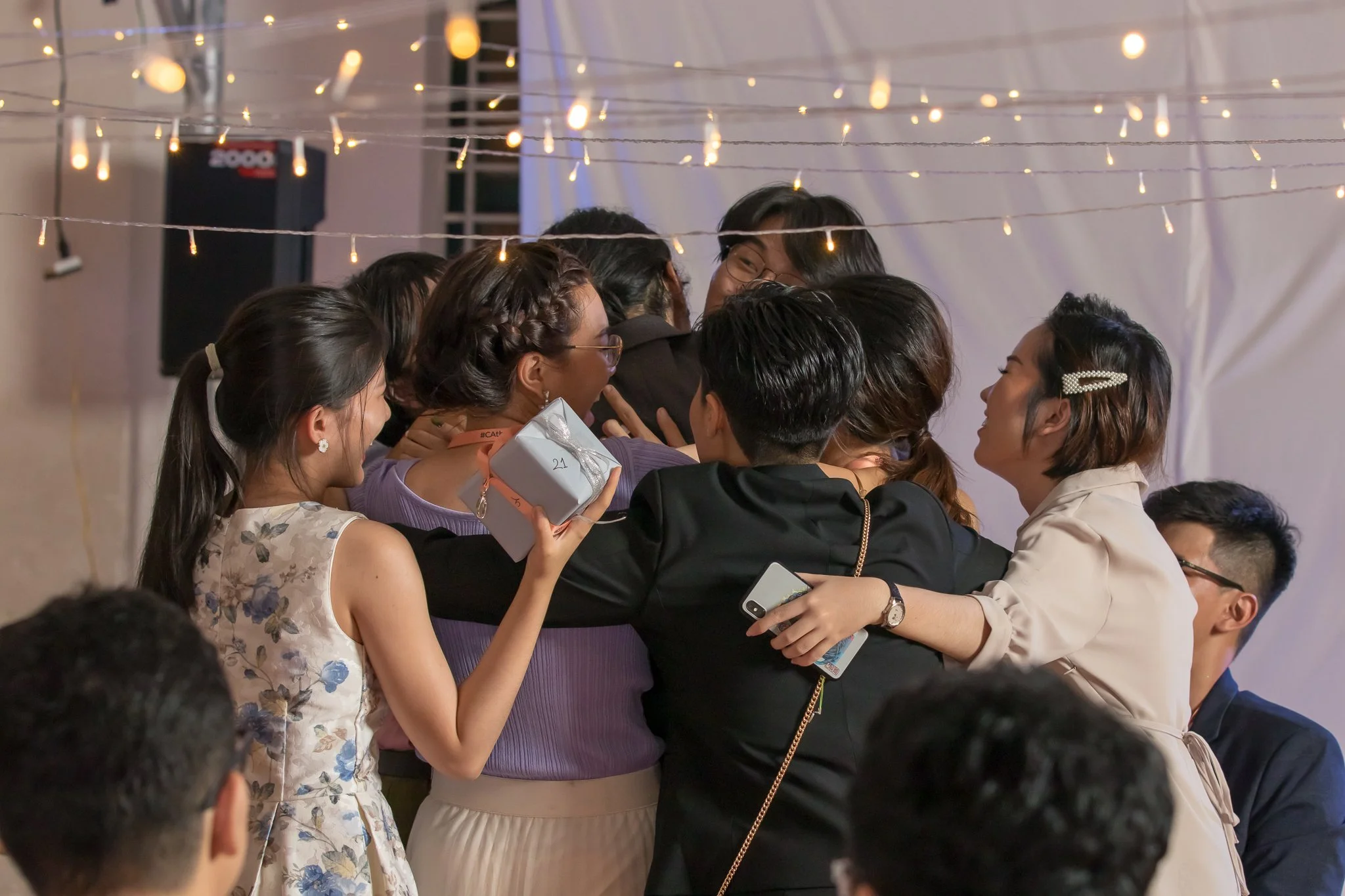 Group of smiling friends hugging each other at a celebration, with string lights overhead.