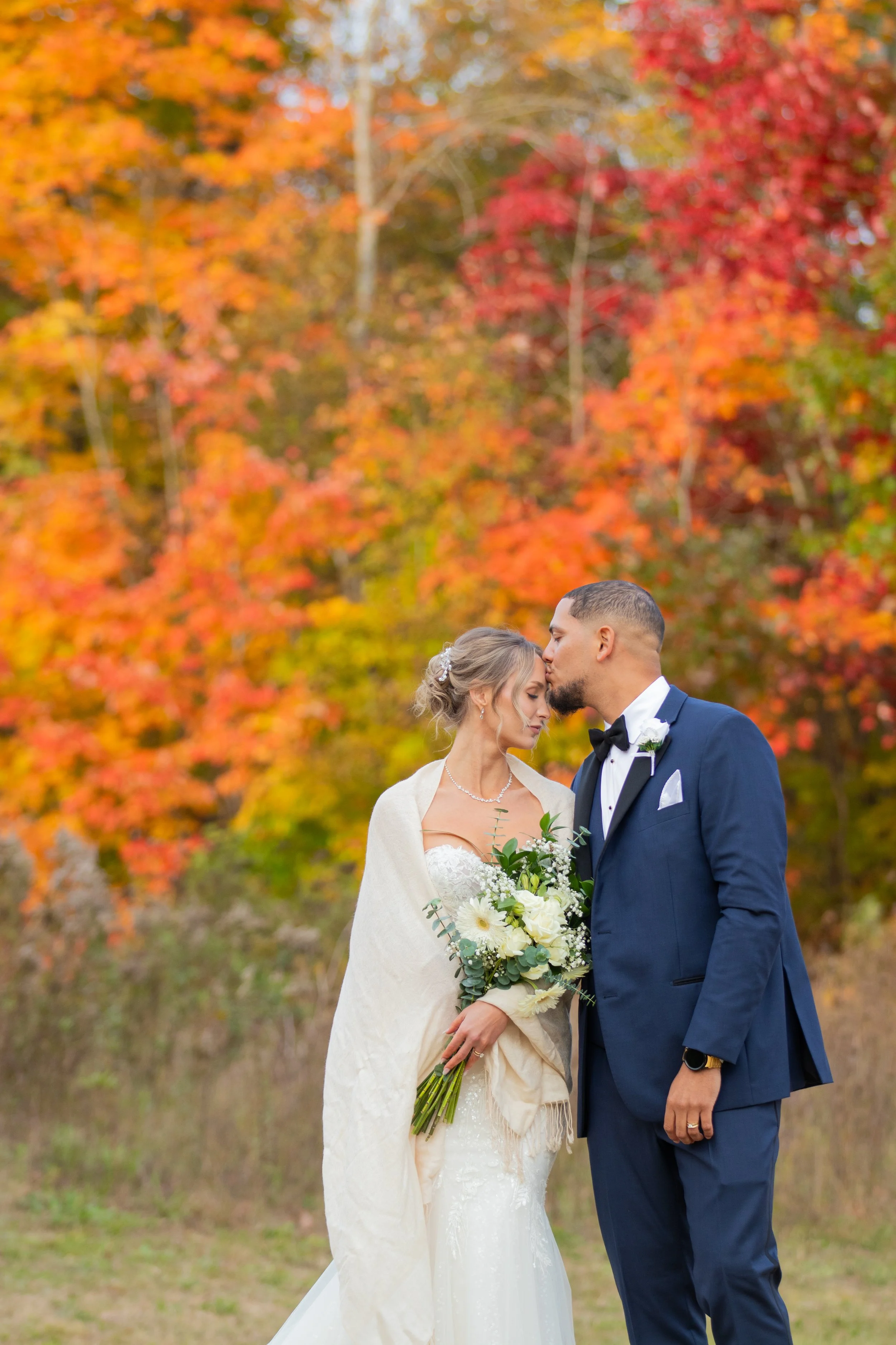A bride and groom sharing a kiss outdoors during autumn, with colorful fall foliage in the background.