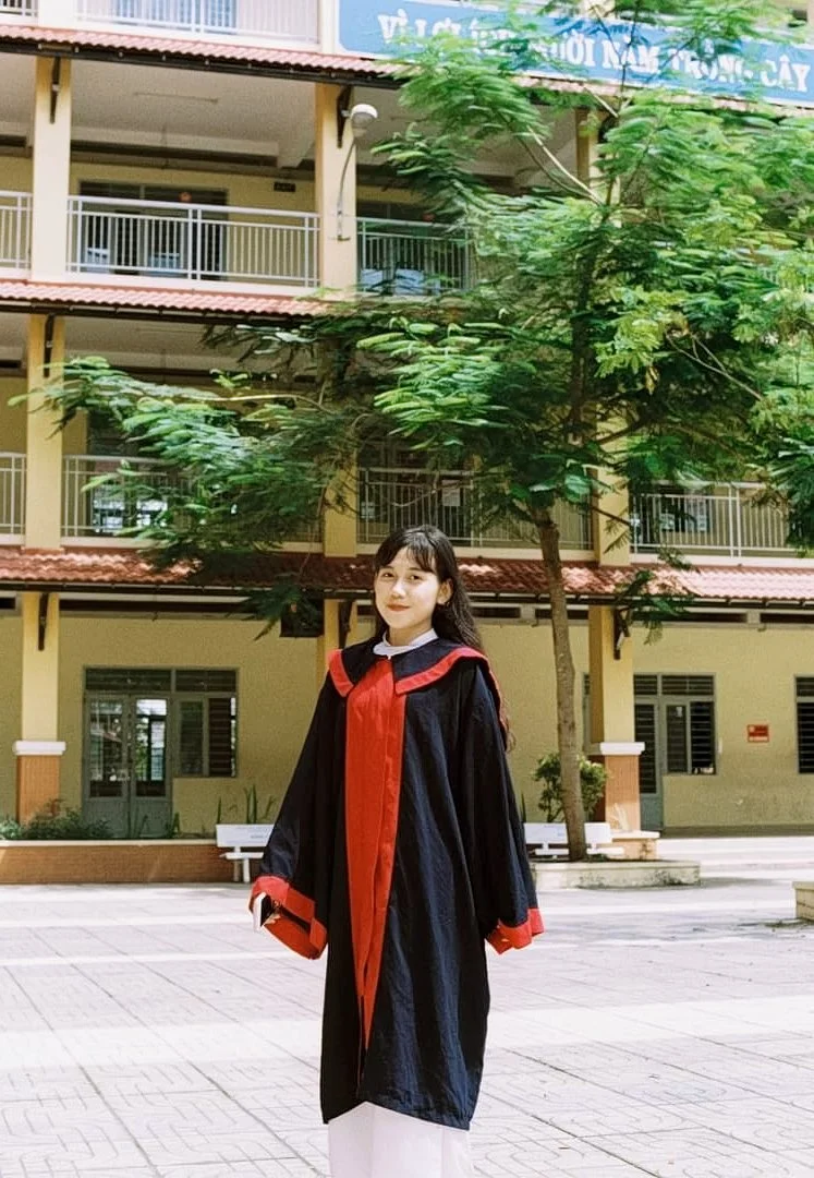 A young woman in a black graduation gown with red accents standing outdoors in front of a multi-story building and trees.
