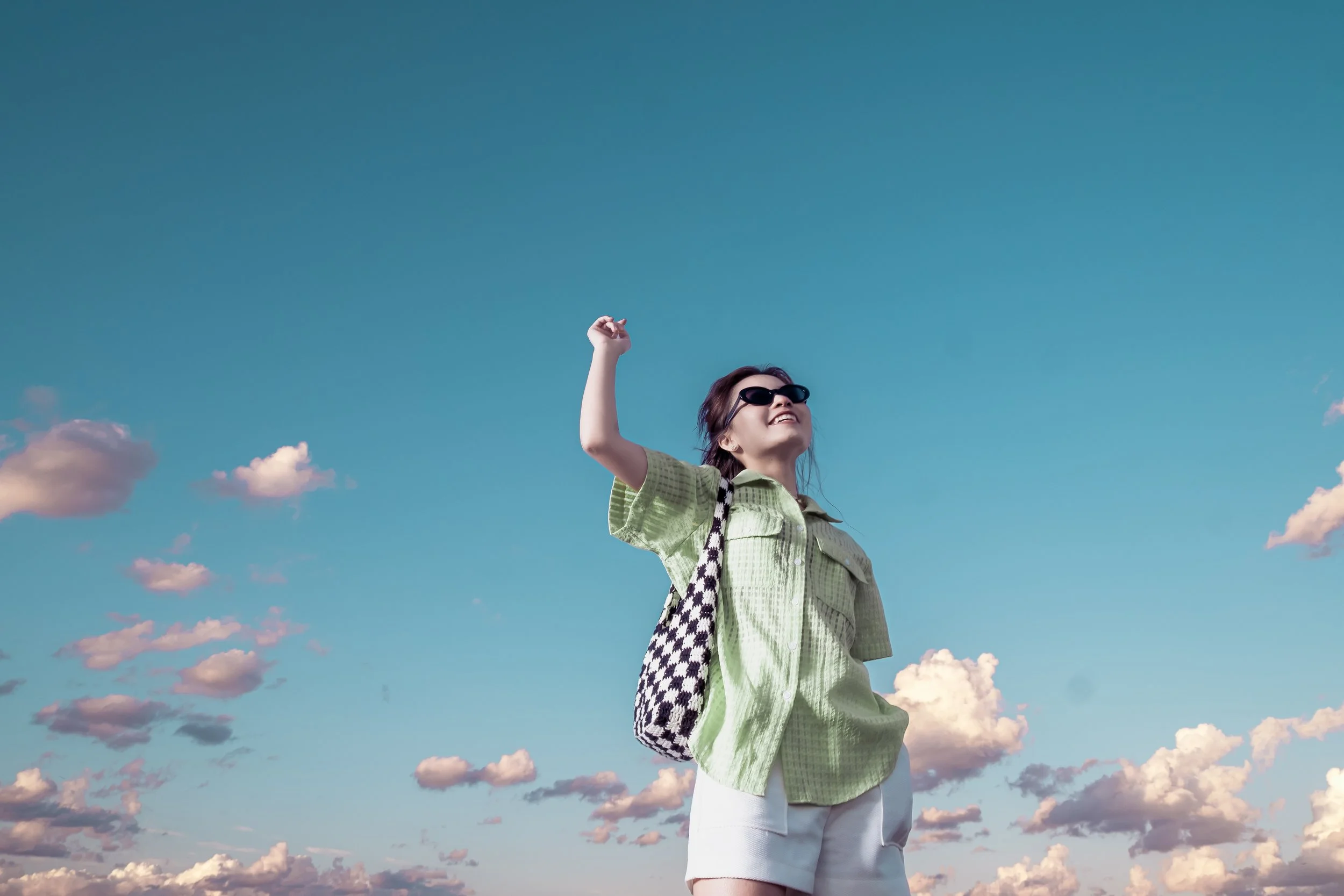 Young woman wearing sunglasses smiling and raising her right arm, standing outdoors against a bright blue sky with scattered clouds, carrying a patterned bag over her shoulder.