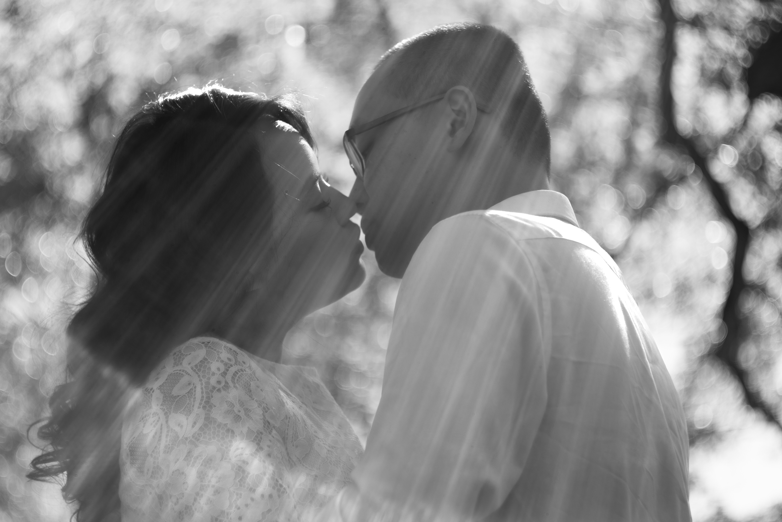 A black and white photo of a couple about to kiss outdoors with sunlight shining through trees in the background.