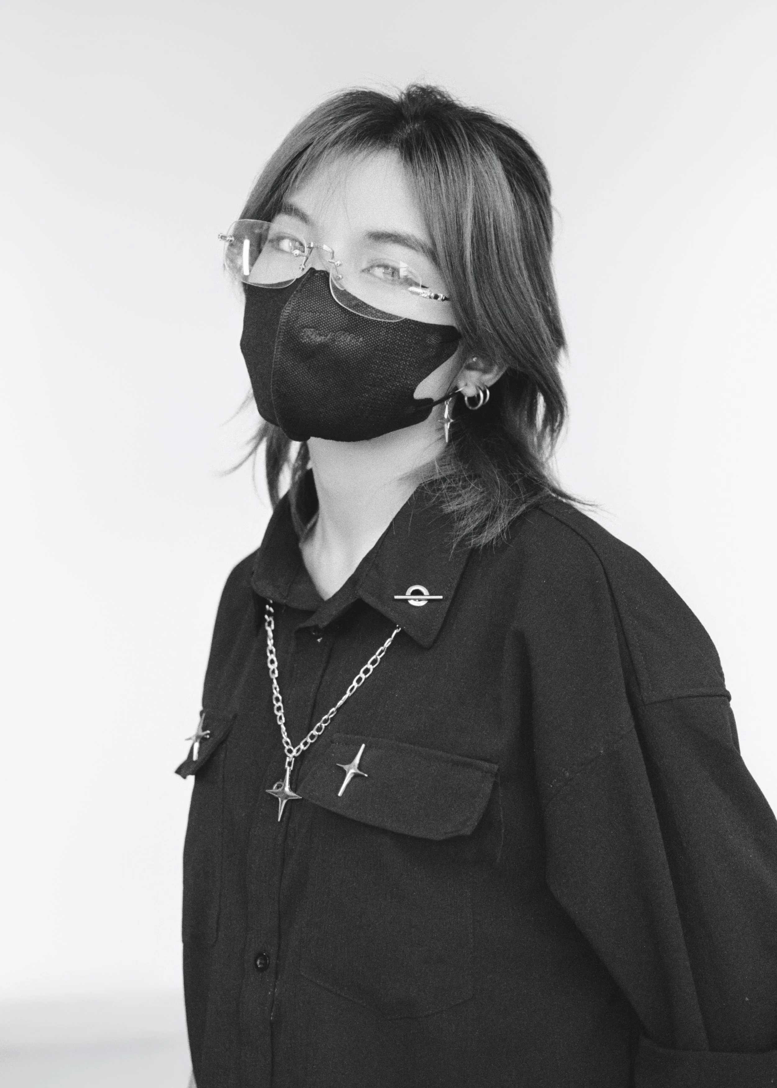 A person with shoulder-length hair, wearing glasses and a black face mask, dressed in a black shirt with silver jewelry, standing against a plain background.