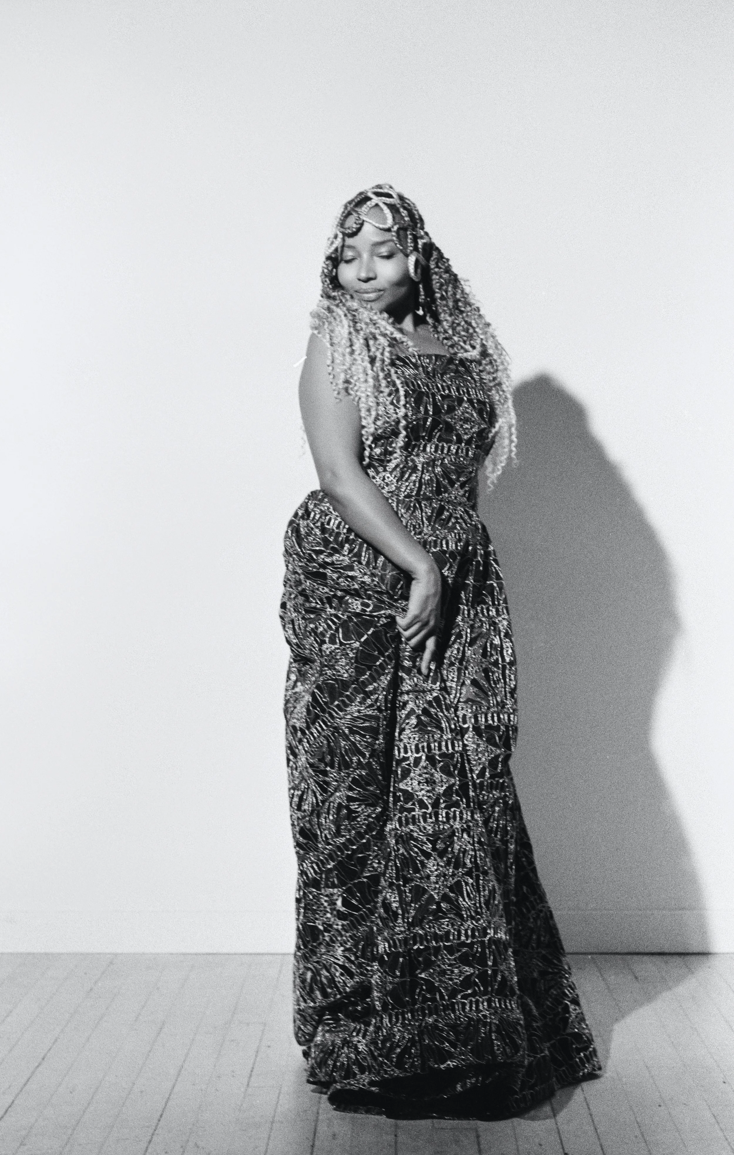 Black and white photo of a woman with long curly hair, wearing a patterned dress, standing against a plain background with her eyes closed and one hand touching her dress.