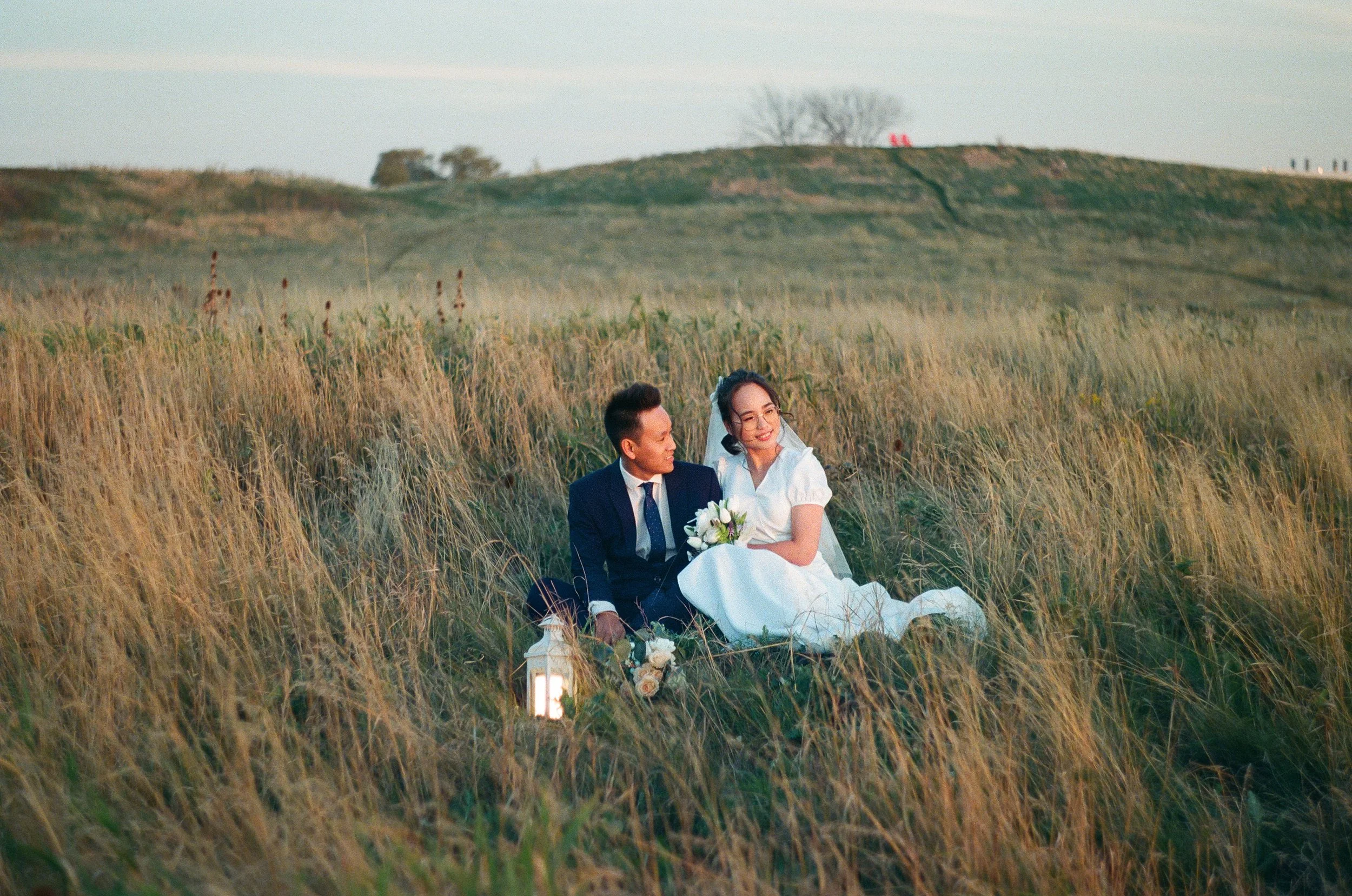 A couple dressed in wedding attire sitting in a field of tall grass during sunset, holding a bouquet and a lantern.