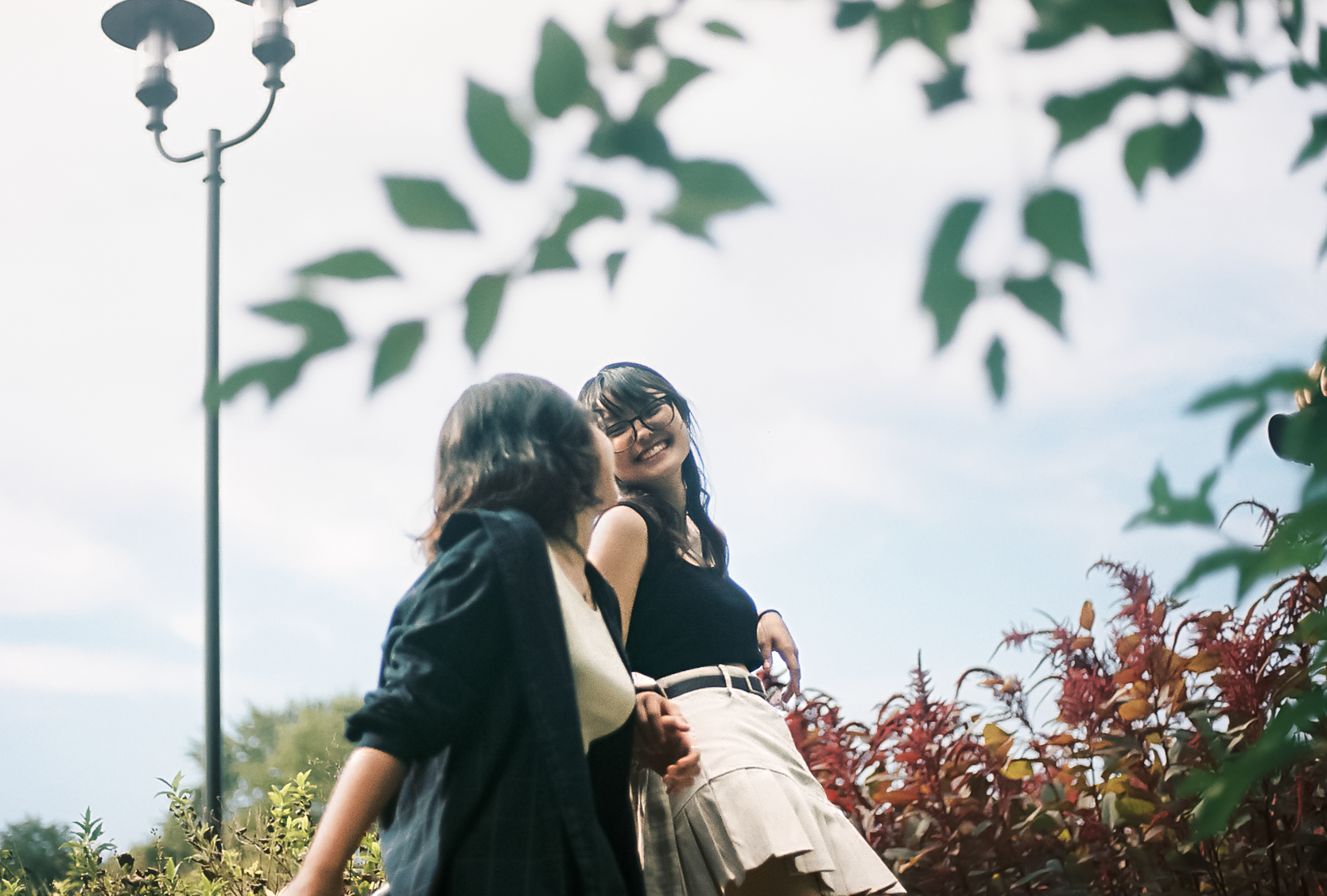 Two young women talking and smiling outdoors among greenery, with one wearing glasses and a black tank top, and the other wearing a dark jacket and lighter top, under a cloudy sky.