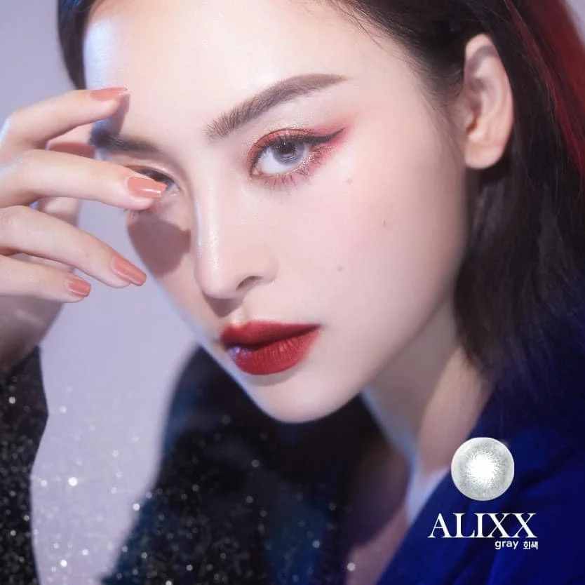 A close-up of a woman with makeup, including red lipstick and eye makeup, touching her eyebrow with her fingers. She has dark hair with red highlights and is wearing a dark outfit with sparkles. The image includes a logo with a sphere and the text "A