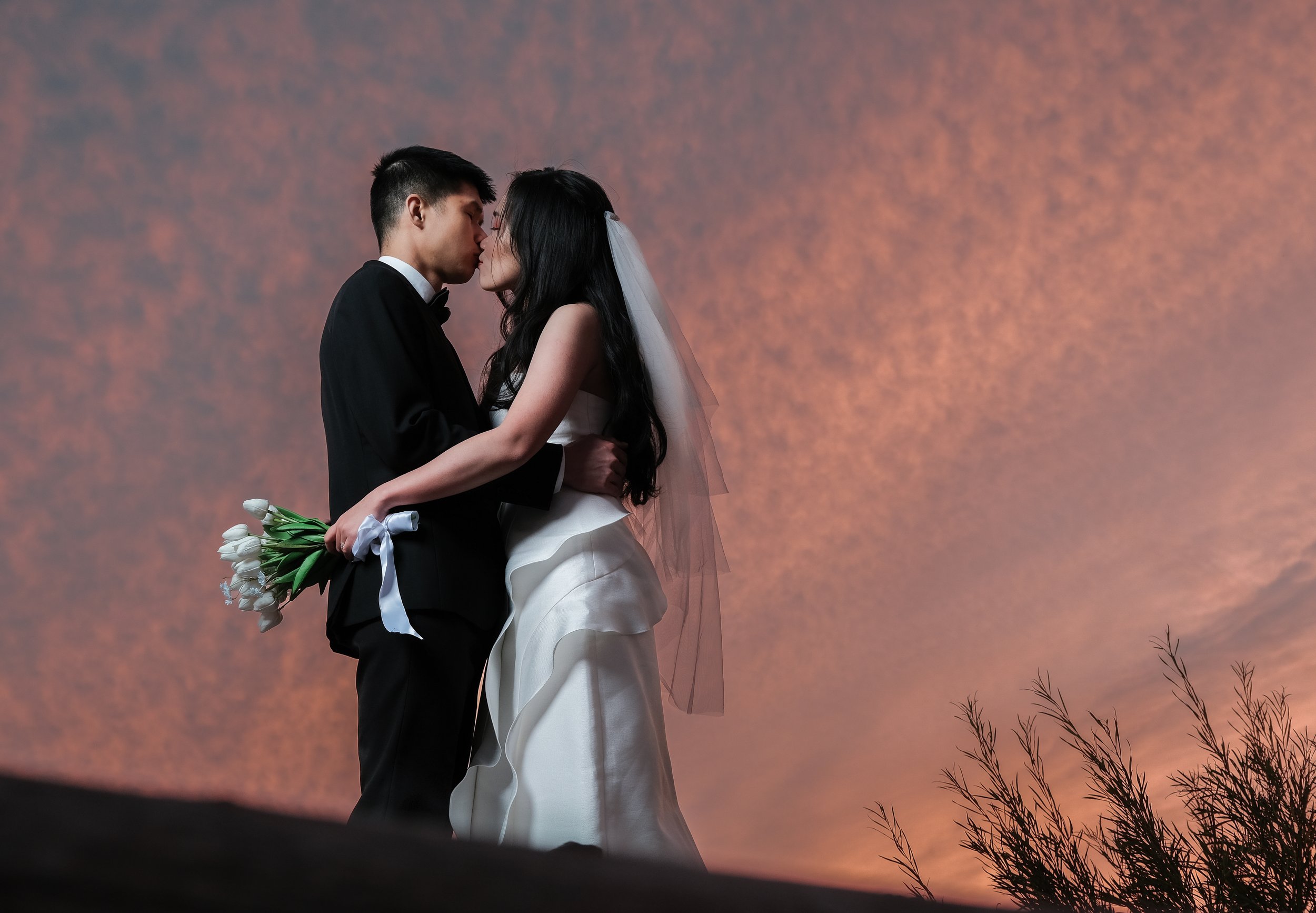 A bride and groom are standing close, about to kiss, with a sunset sky in the background. The groom is holding a bouquet of white tulips behind his back.
