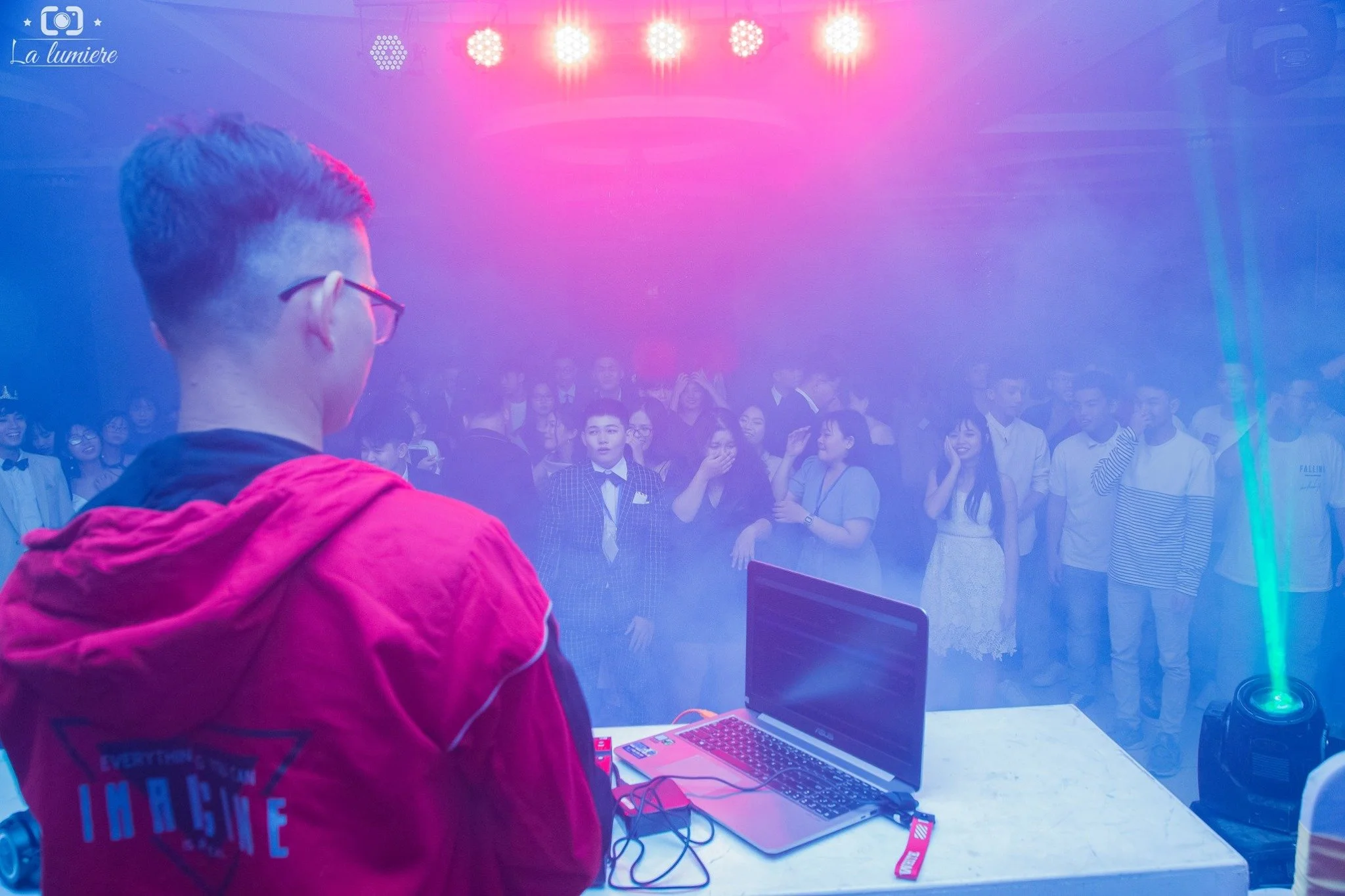A DJ wearing glasses and a red jacket performs at a nightclub with a crowd of young people dancing and enjoying the music. Colorful lights illuminate the scene, and a laptop is on the DJ's table.