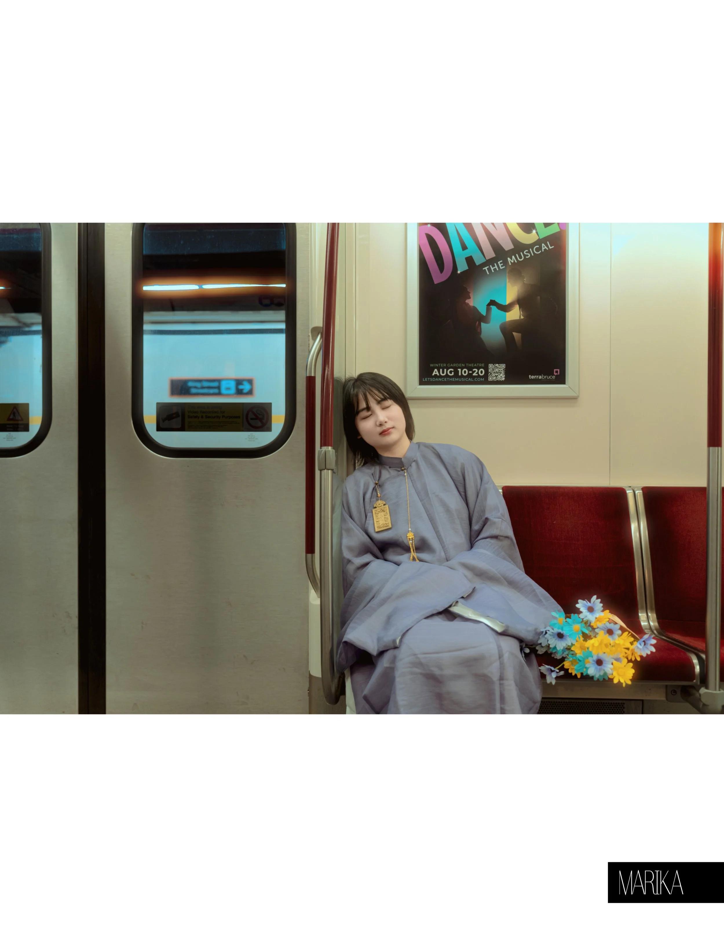 A young woman in a gray traditional dress sitting on a subway train with her eyes closed. She has a bouquet of colorful paper flowers on her lap and a gold pendant around her neck. Behind her is a poster for the musical 'Dance the Musical' and a wind