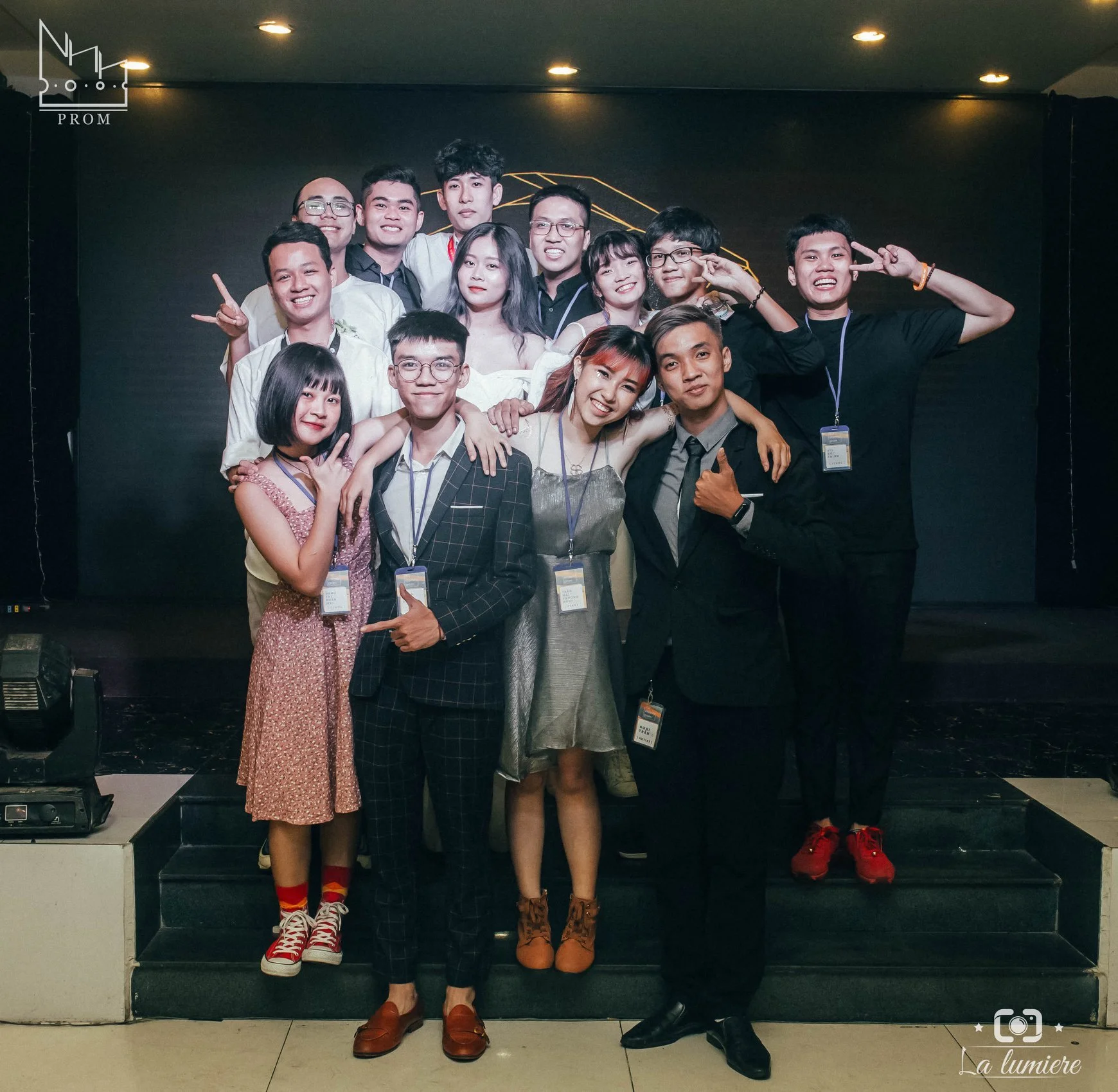 Group of 12 young adults at a formal event, standing on stairs in front of a black backdrop, smiling and posing for the camera.