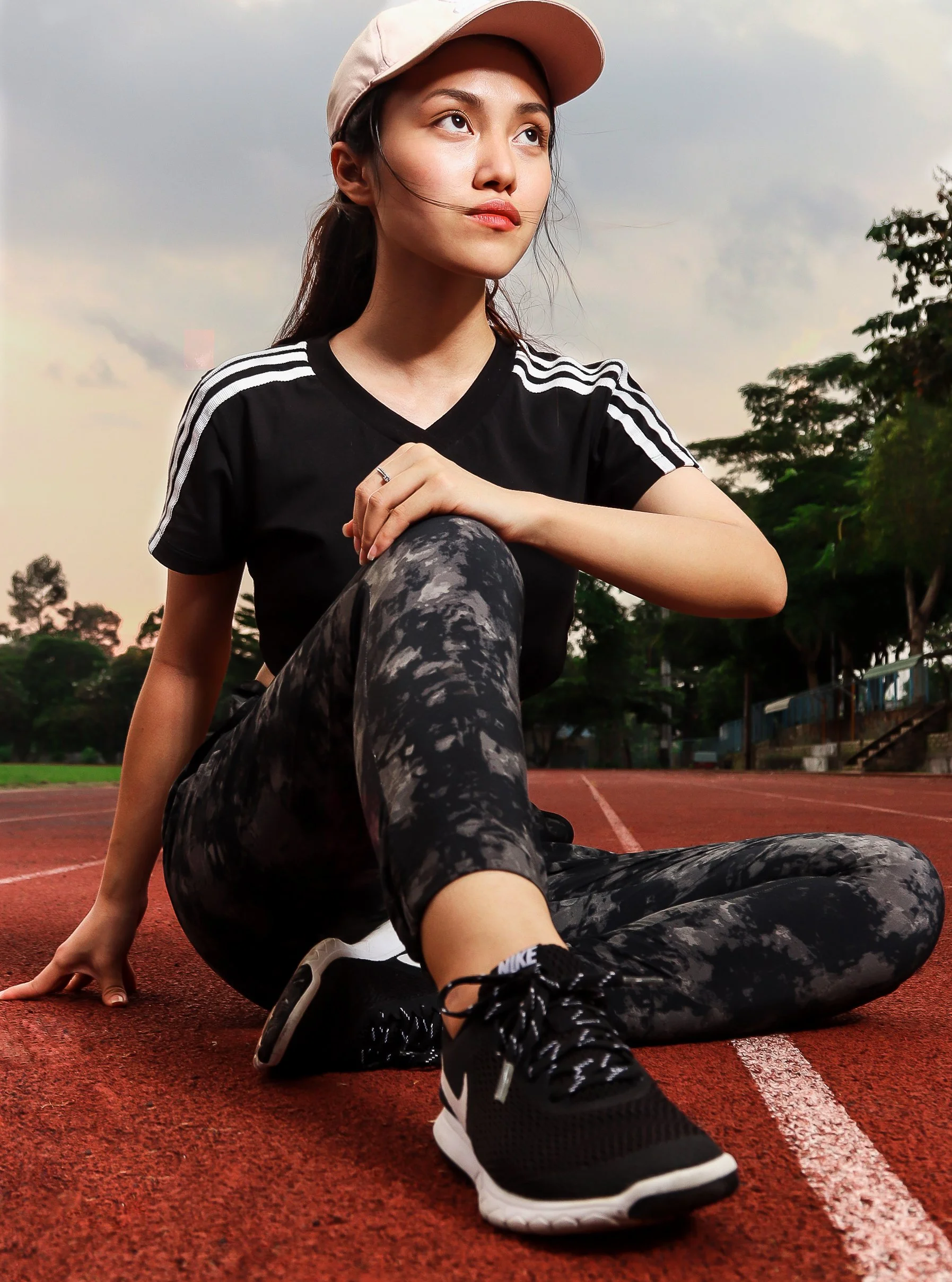 Young woman sitting on a red running track, wearing a black and white athletic outfit, black Nike running shoes, beige cap, and looking thoughtully into the distance. Trees and a cloudy sky are in the background.