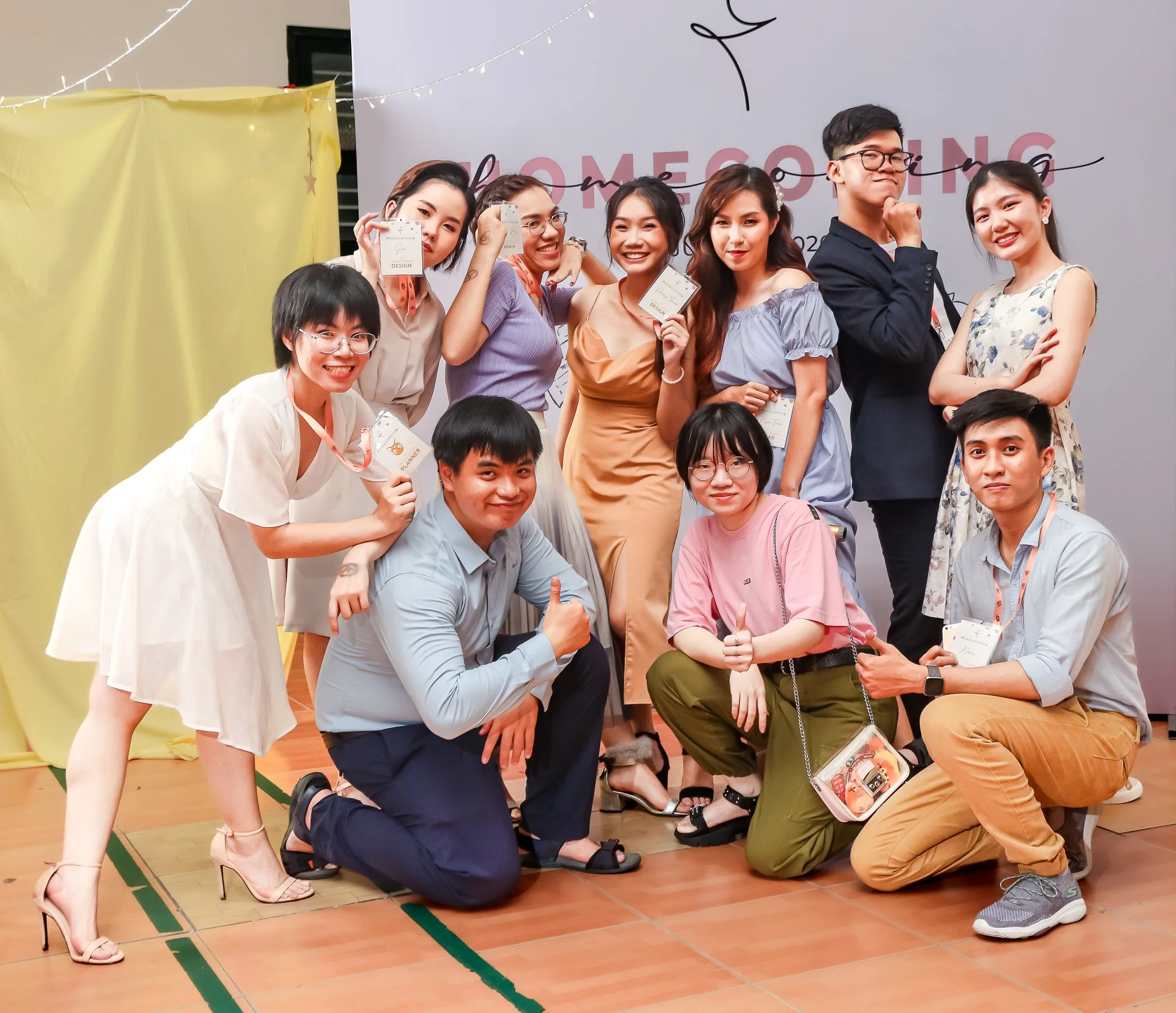 Group of nine young adults at a celebration event, some holding awards or cards, smiling and posing for the photo in front of a decorated wall with the partially visible word 'Welcome'.