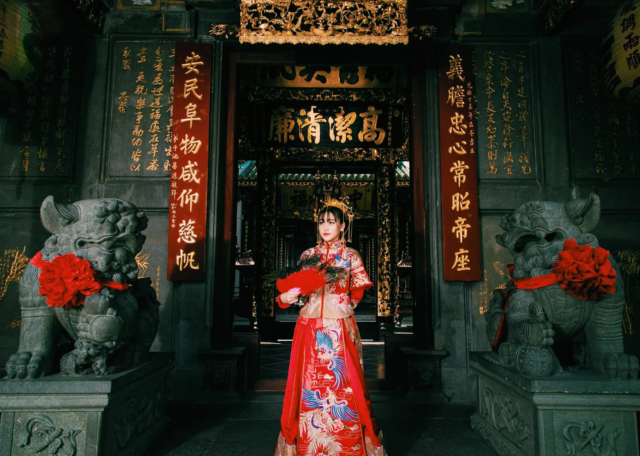 A woman in traditional Chinese attire standing in front of an ornate, decorated temple entrance, with Chinese calligraphy on red and black plaques, stone guardian lion statues with red ribbons, and intricate gold detailing.