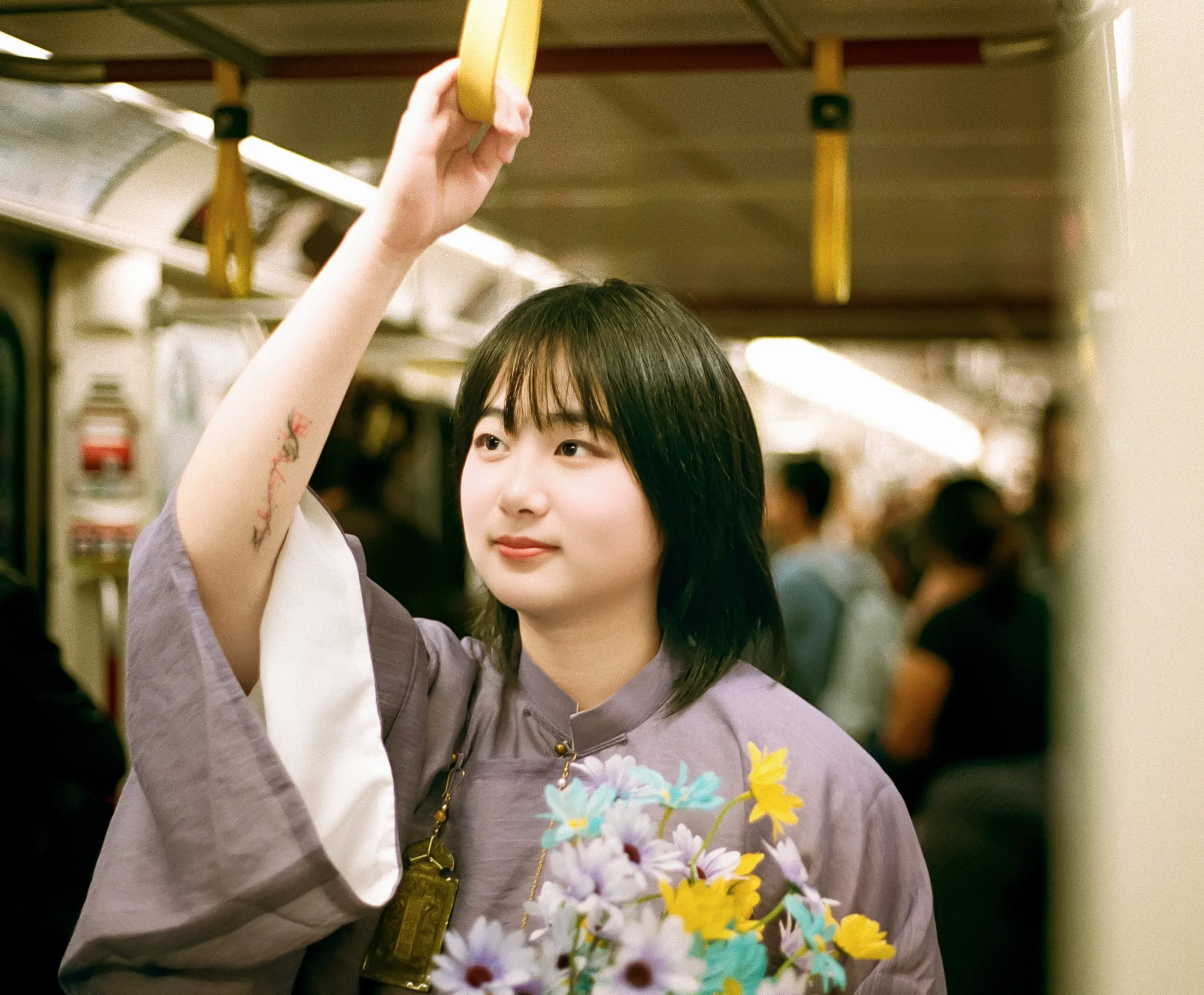Young woman with black hair holding a bouquet of colorful flowers and standing on a subway train, holding a yellow handrail.
