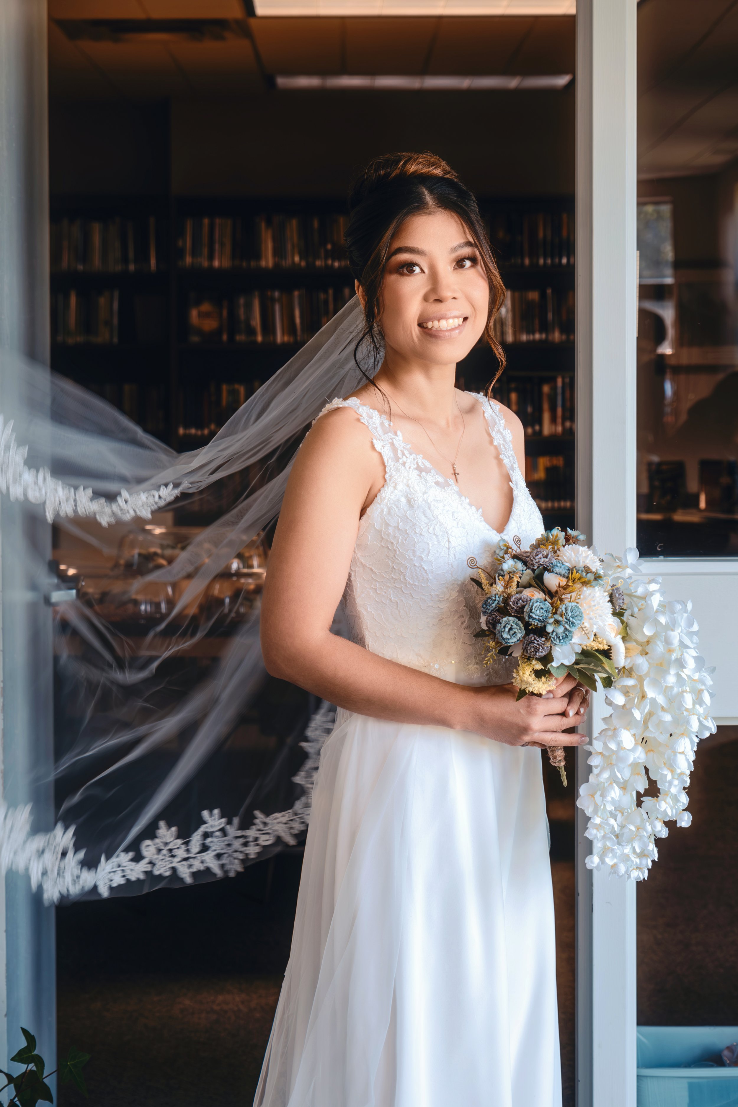 A bride with dark hair styled in an updo, wearing a white lace wedding dress and a veil, standing indoors and holding a bouquet of white and pastel flowers with greenery, smiling at the camera.