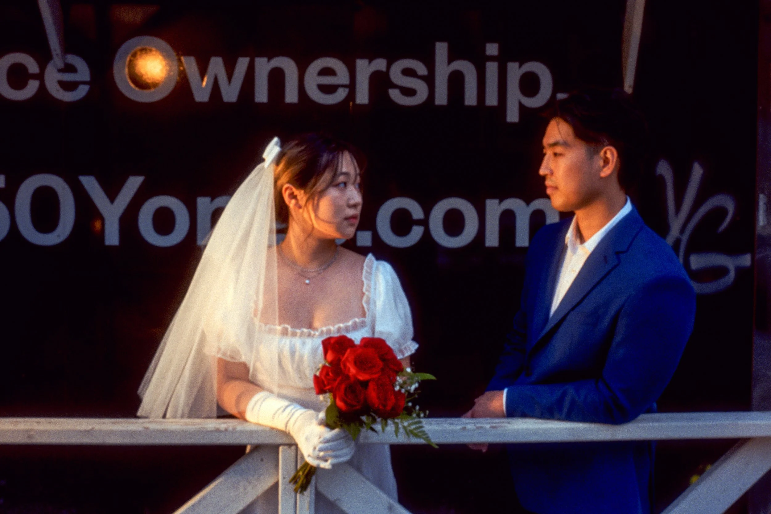 A bride in a white dress and veil holding a bouquet of red roses, looking at a groom in a blue suit, on a bridge at sunset.