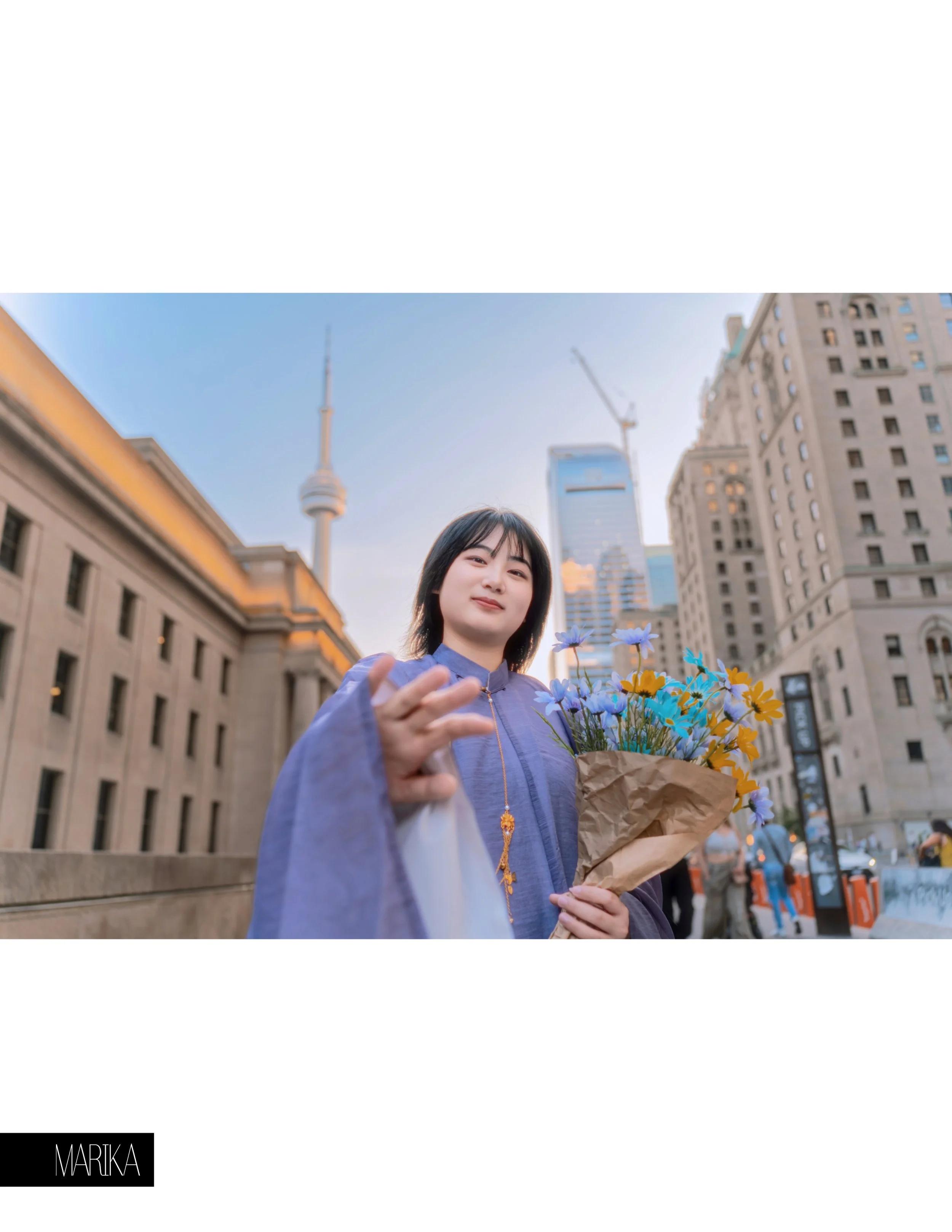 A young woman dressed in traditional clothing holding a bouquet of yellow and blue flowers in an urban cityscape with tall buildings and a clear sky.
