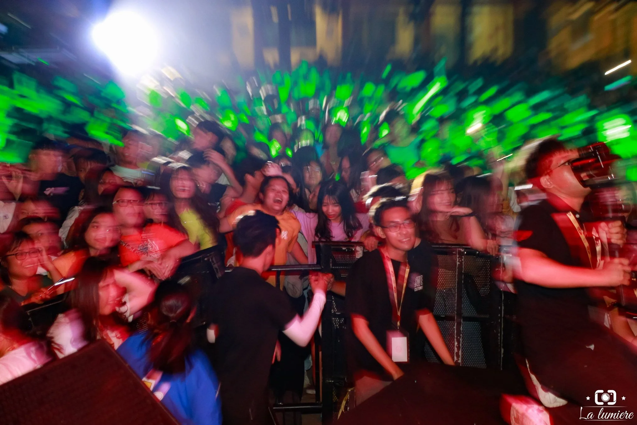 Crowd of people at a lively event with green lights and a bright white spotlight, showing excitement and cheer.