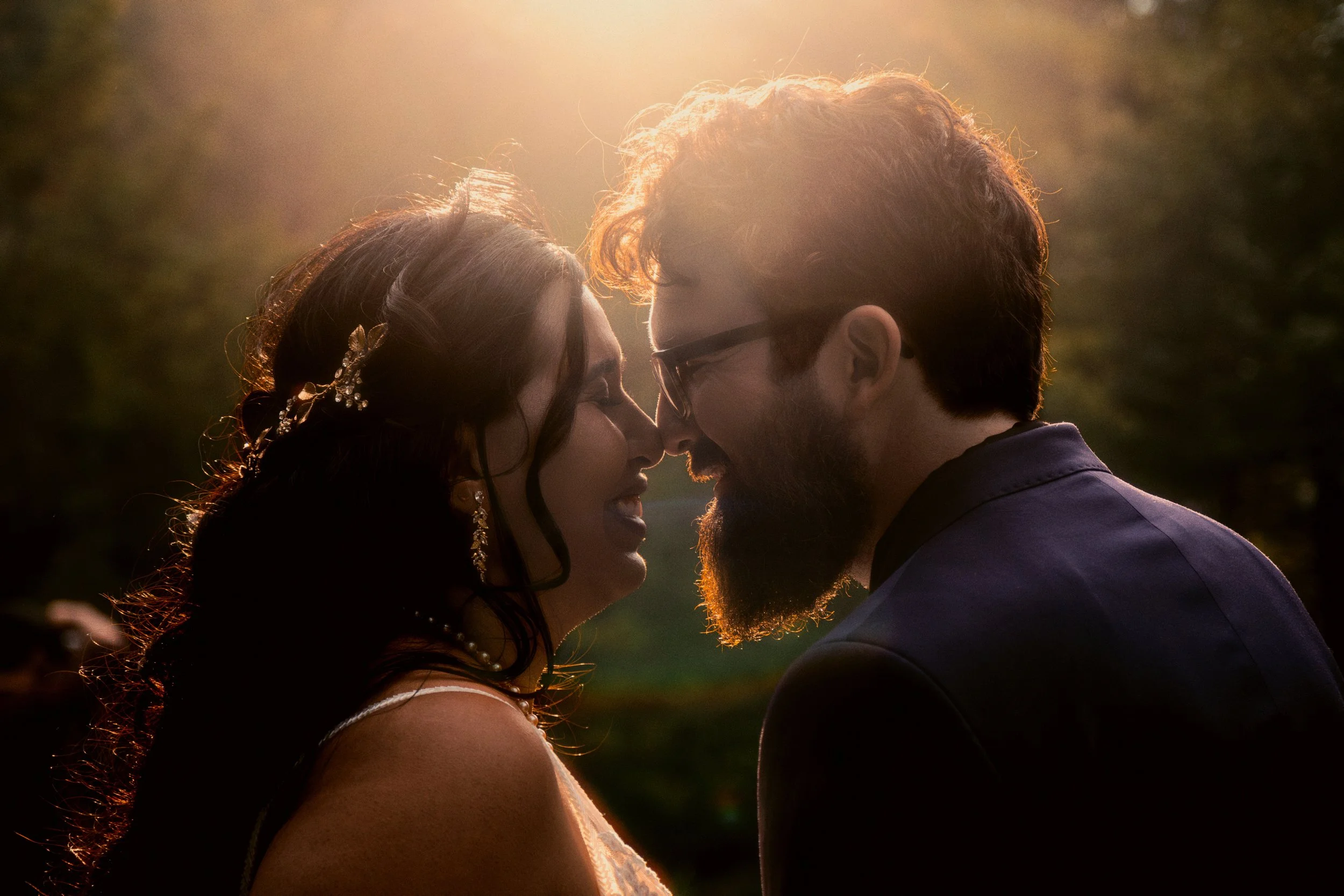 A couple touching foreheads at sunset, outdoors, with the woman wearing wedding attire and jewelry and the man in a suit, backlit by the sun.