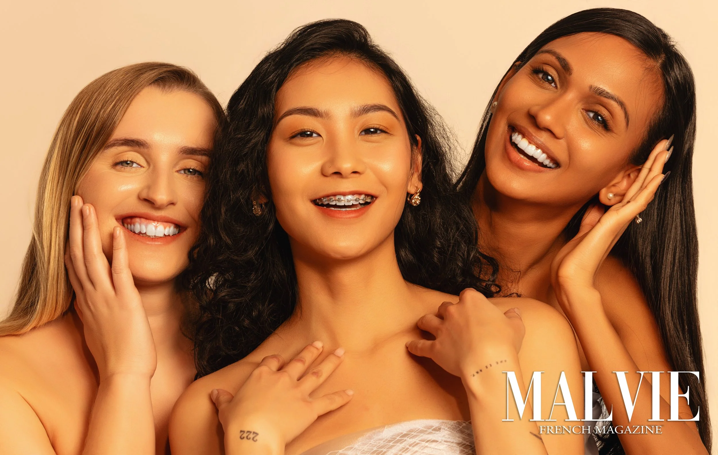 Three smiling women with diverse appearances, wearing minimal clothing, posing closely together for a photo against a light background. The magazine cover text reads "MALVIE FRENCH MAGAZINE."