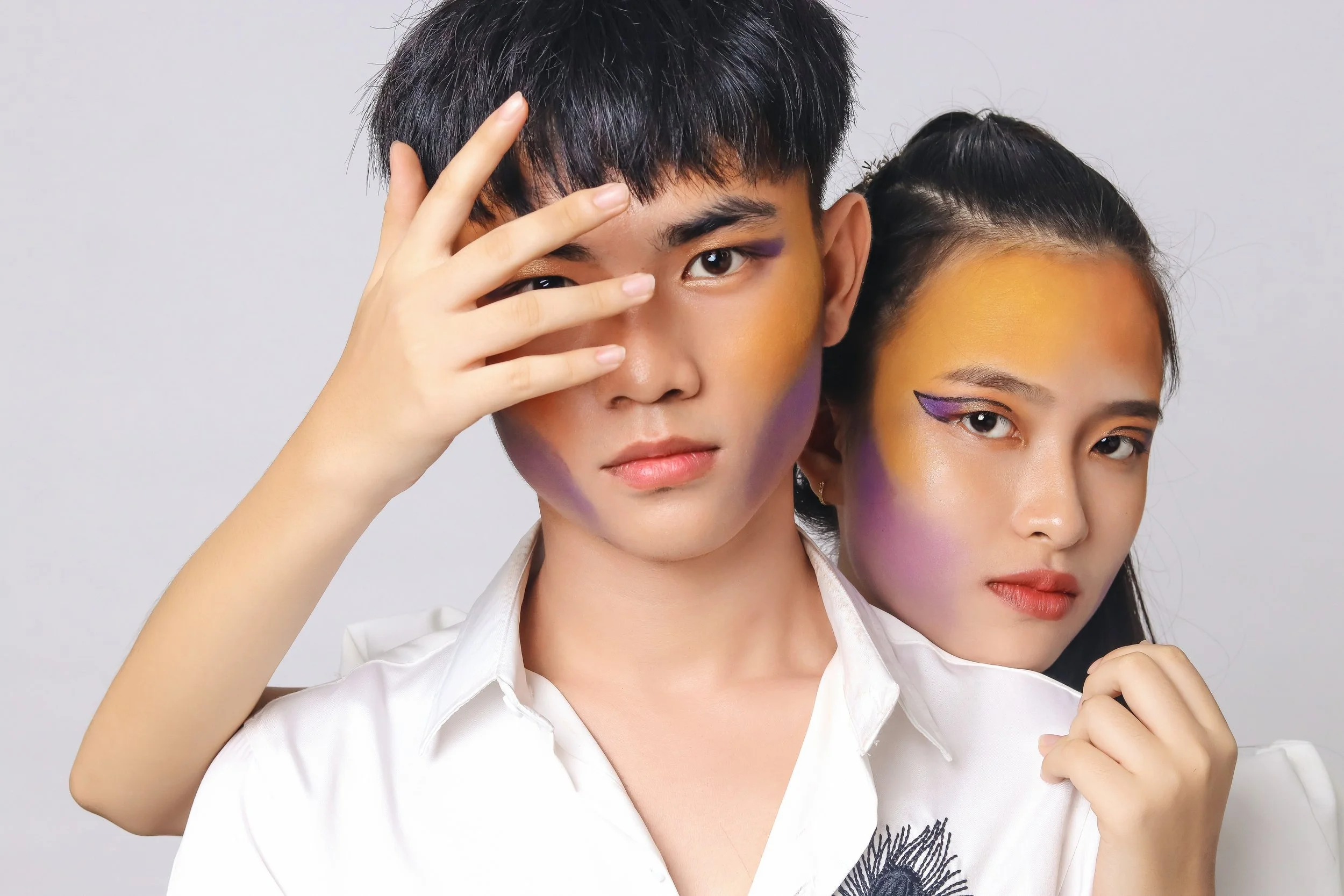 Two young individuals with bold, colorful makeup on their faces, posing closely against a plain white background.