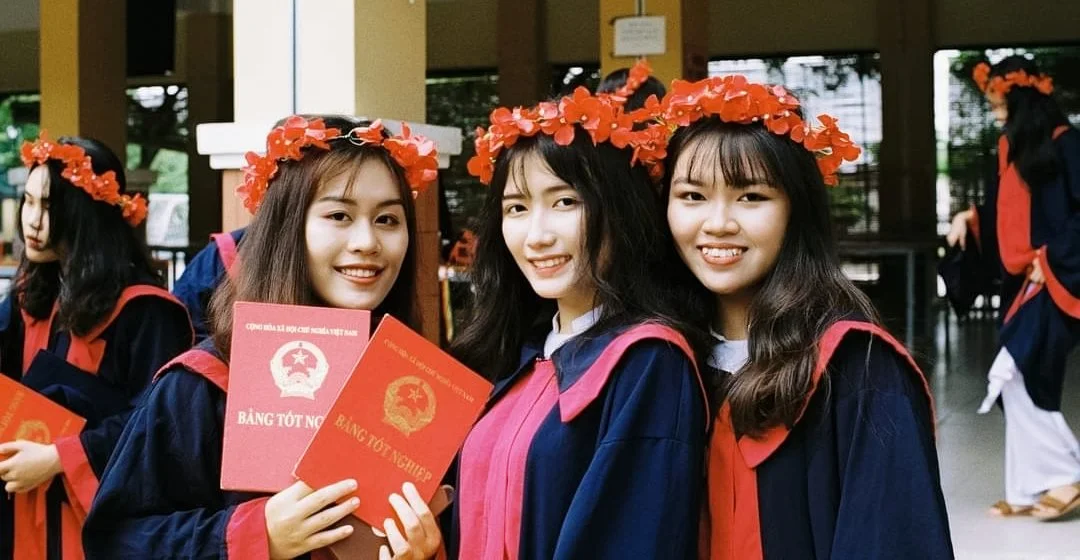 Three young women in graduation gowns with red and pink accents and red flower crowns, holding red diplomas, smiling for the camera.
