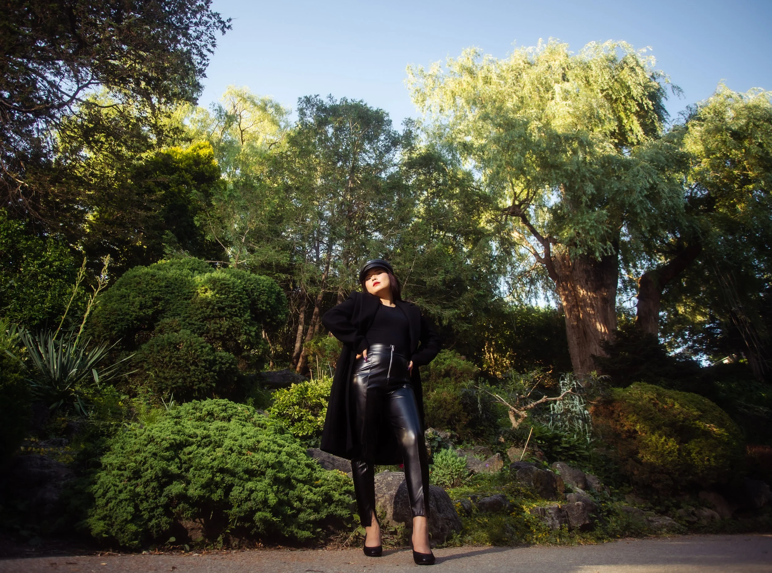 A woman dressed in black leather pants, black coat, and black heels standing outdoors on a paved path with green trees and bushes in the background.