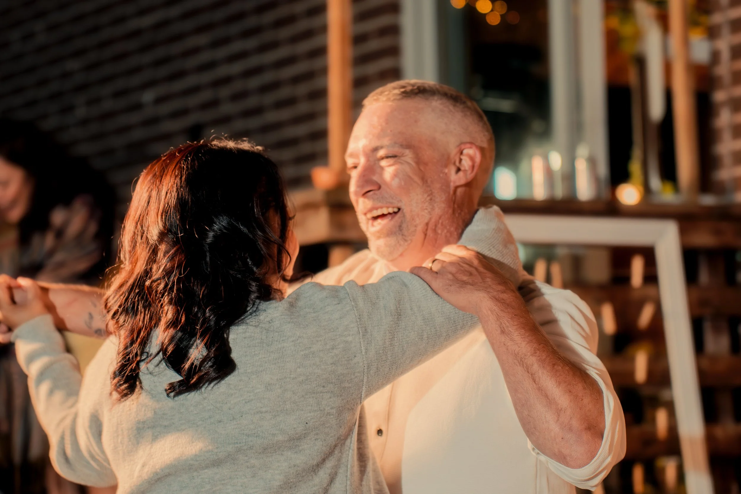 A man and a woman dancing together, smiling and embracing each other in a warm, cozy setting with a brick wall and warm lighting.