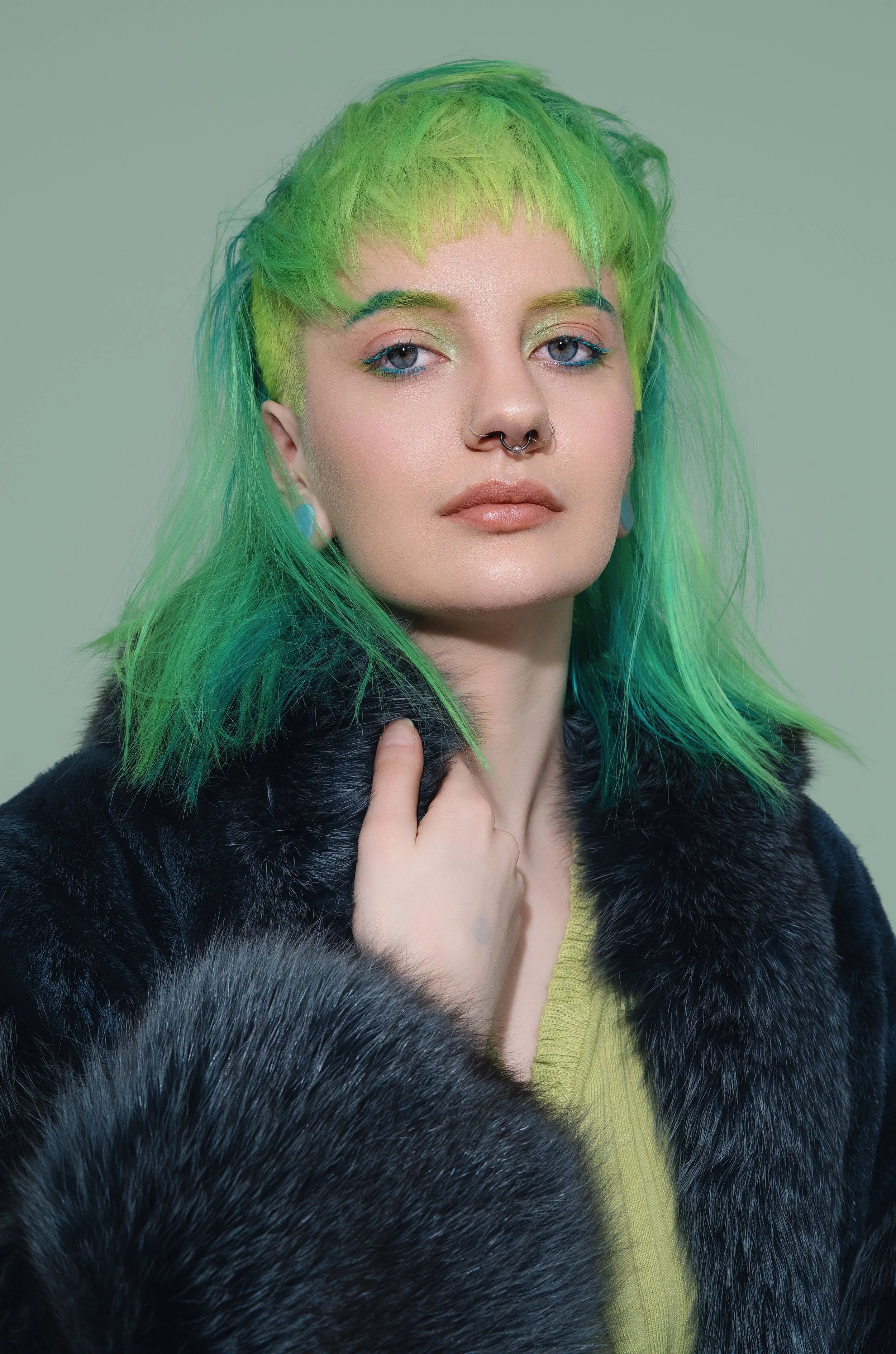 Portrait of a woman with neon green hair, wearing a black fur coat and a yellow top, with a neutral background.