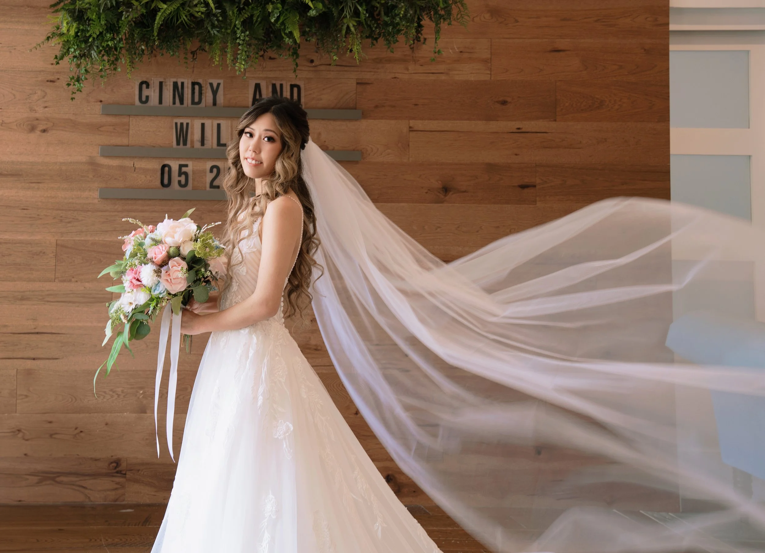 A bride in a white wedding dress and veil holding a bouquet of pink and white flowers, standing in front of a wooden wall with a black letter board displaying her name 'Cindy and WIll' and the date '05 2'.
