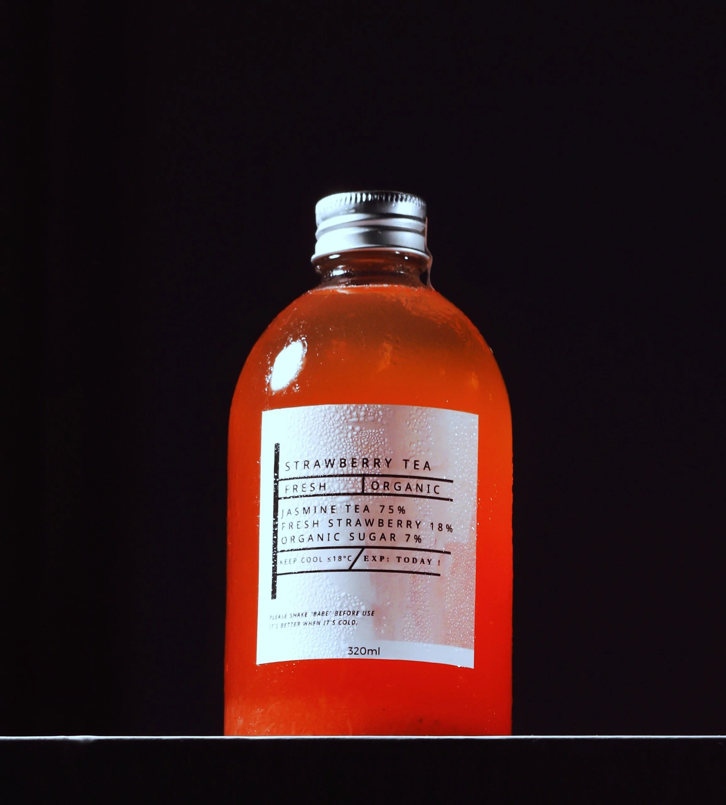 A 320ml orange bottle of strawberry tea with condensation, silver cap, standing on a surface against a black background. The label indicates it is organic, with 75% jasmine tea, 18% fresh strawberry, and 7% organic sugar.