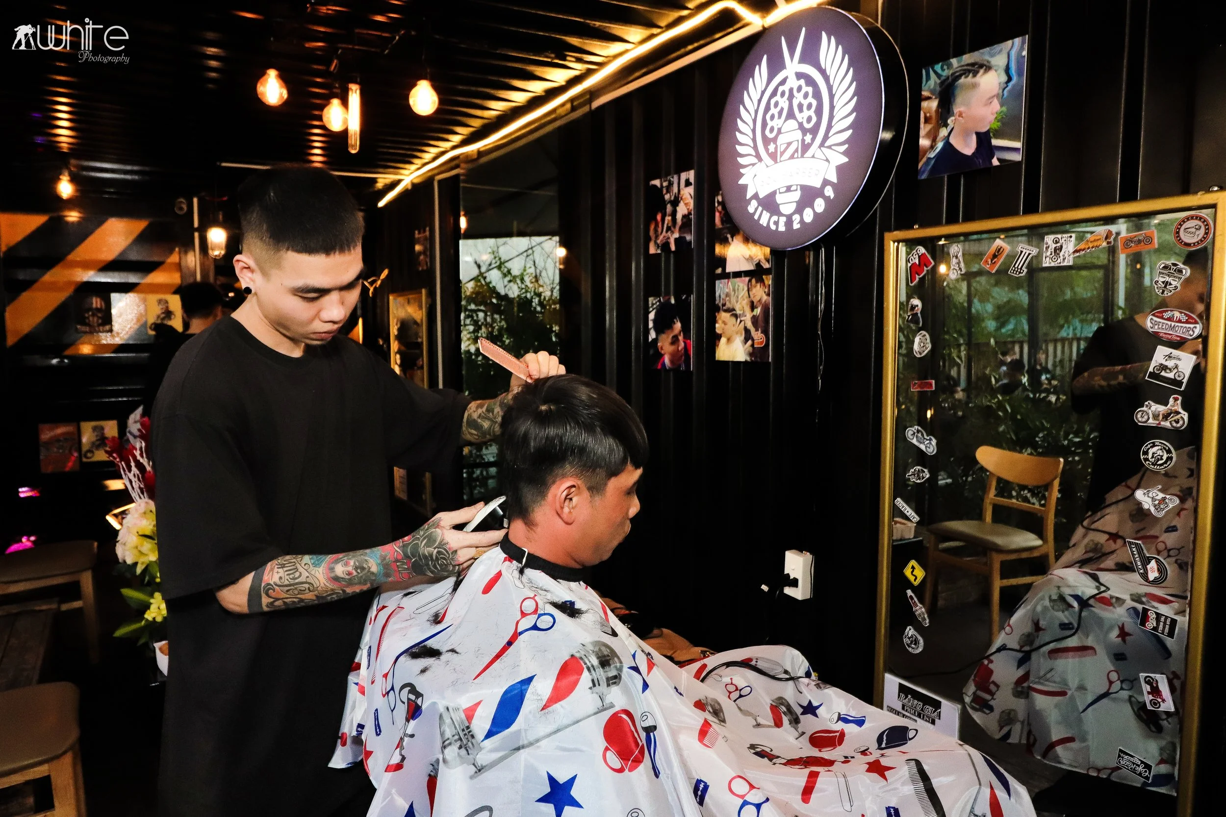 A man getting a haircut at a barber salon, with a hairstylist standing behind him and using a smartphone. The salon has a black interior with wall decorations, photographs, stickers, and a large mirror.
