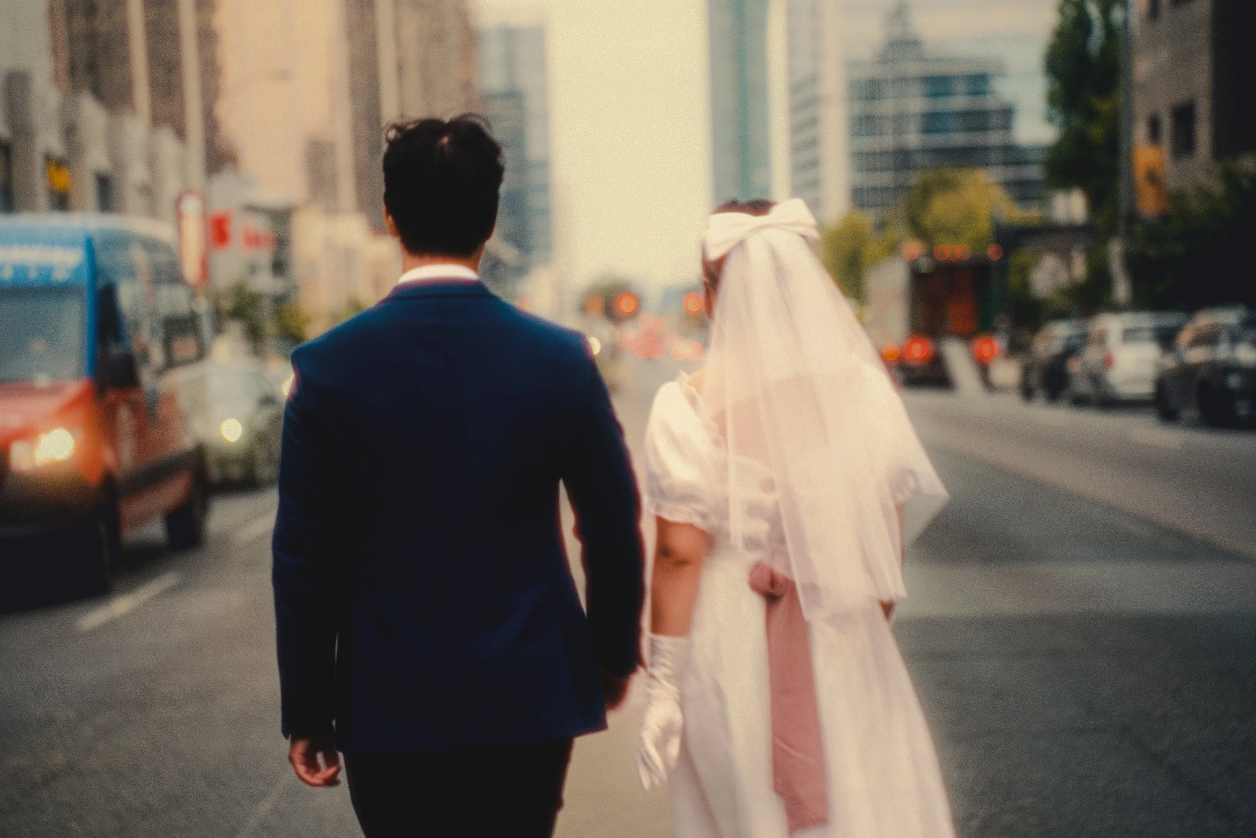 A man and a woman dressed in wedding attire walking down a city street at sunset, with buildings and cars in the background.