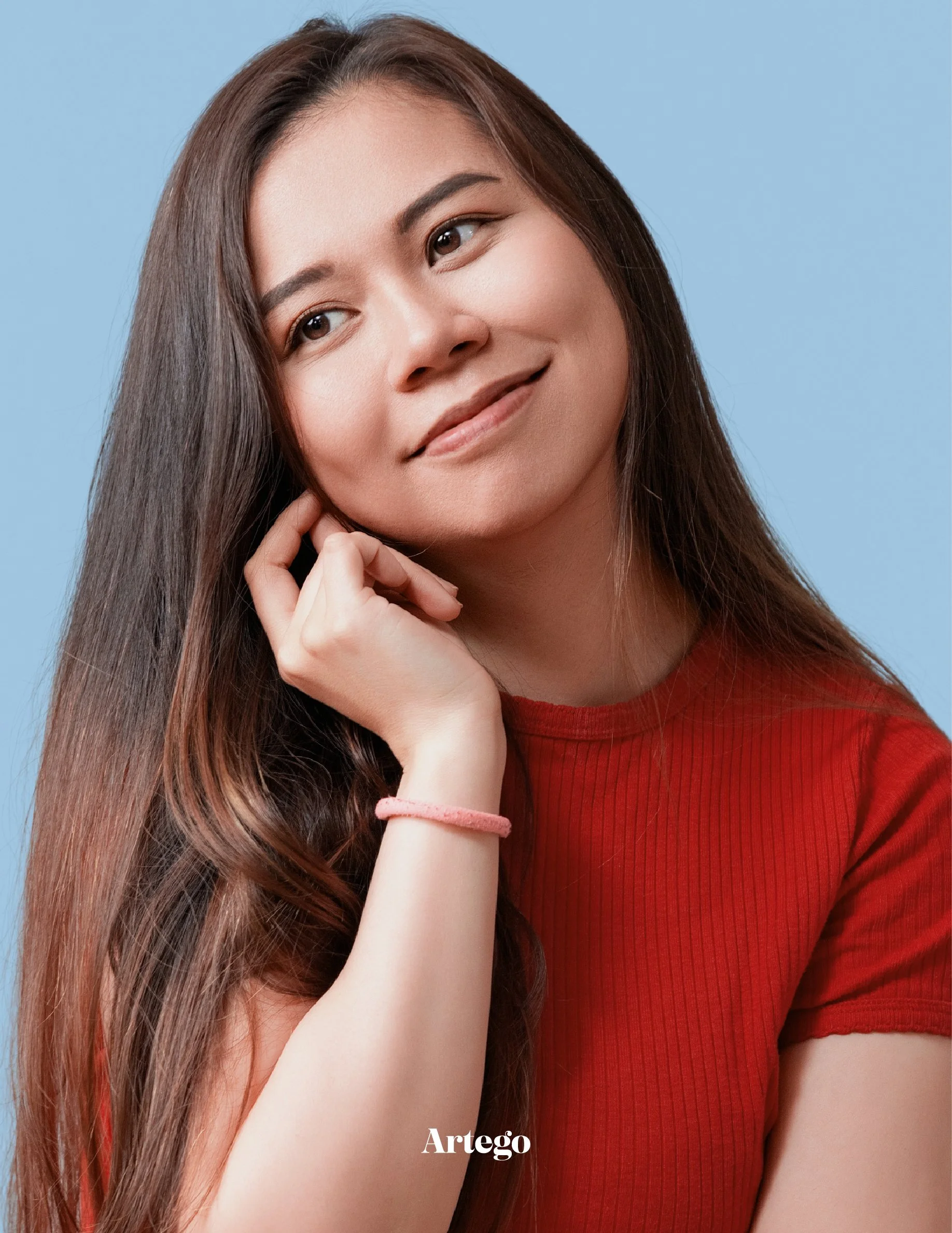 A young woman with long brown hair wearing a red top, posing against a blue background, touching her face with a gentle smile.
