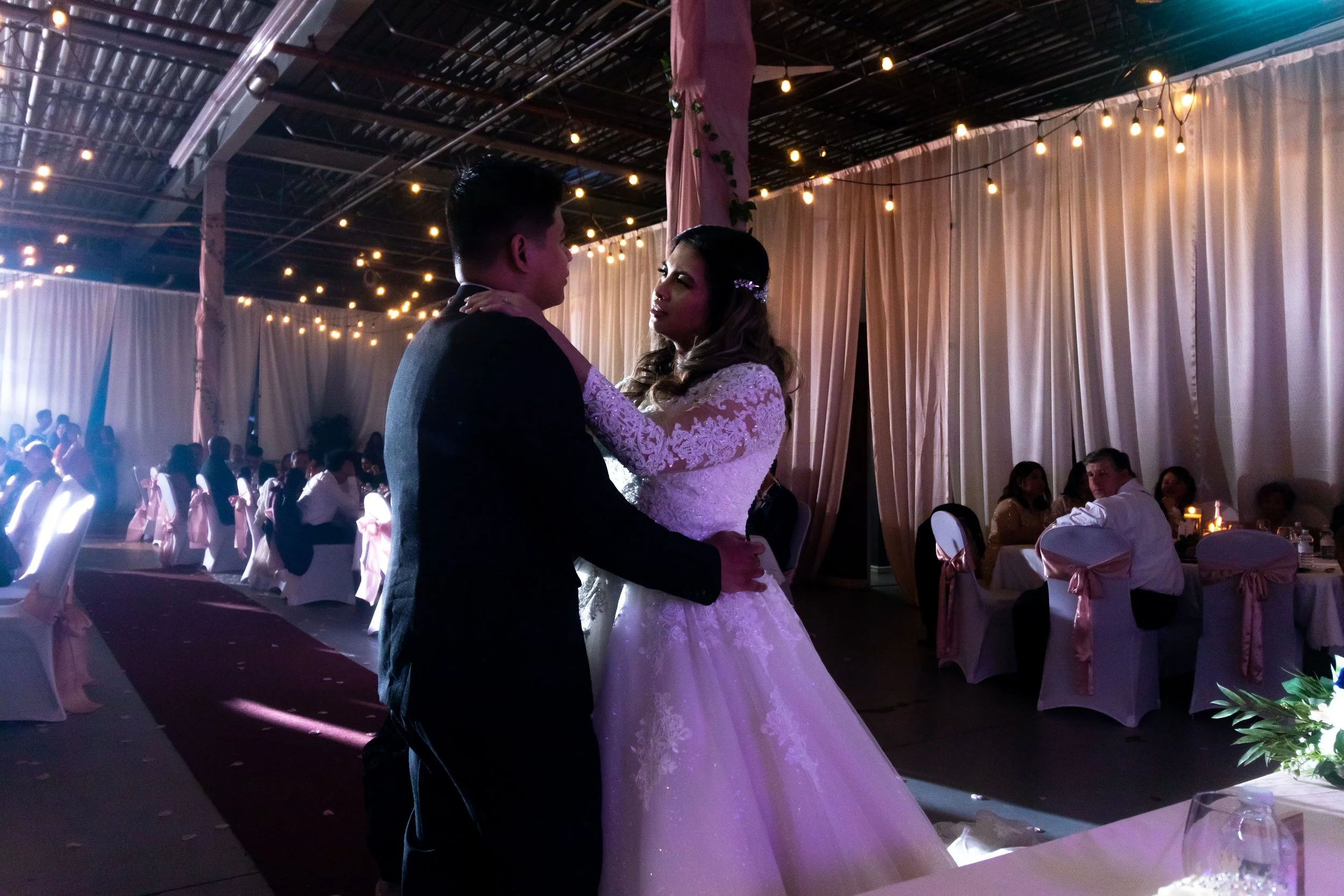 A bride and groom sharing a dance at their wedding reception, illuminated by soft lighting with guests seated in the background.