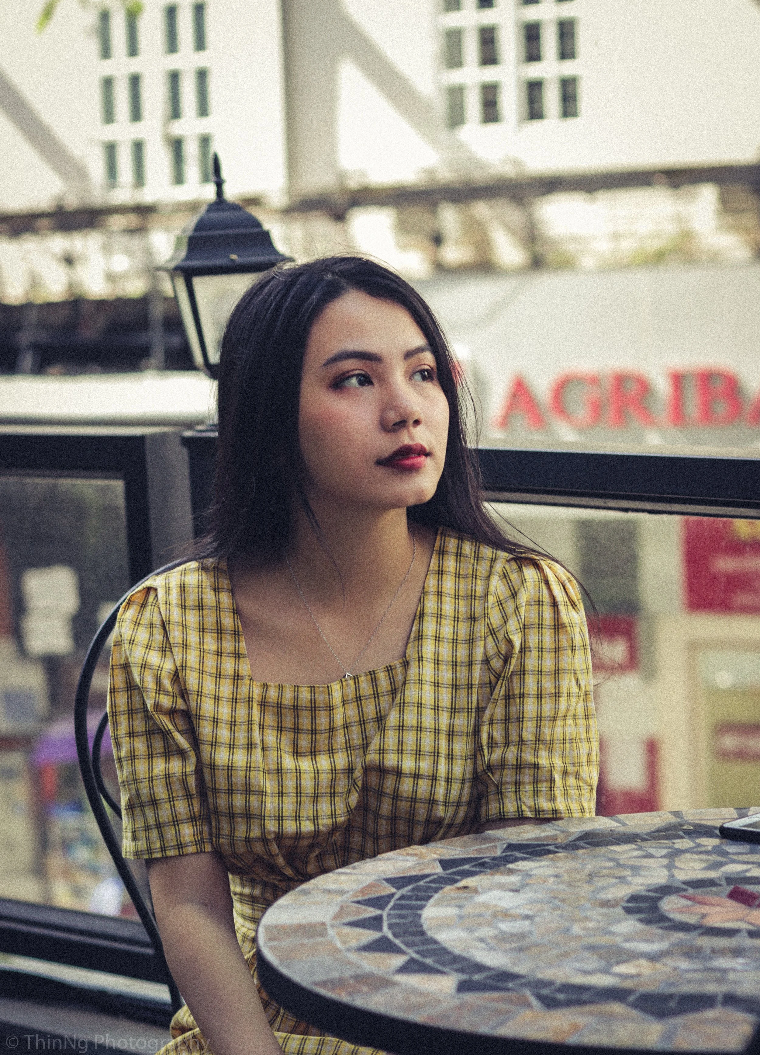 A young woman with long dark hair and lipstick wearing a yellow checkered dress sitting at a mosaic table in a cafe with outdoor greenery and buildings in the background.
