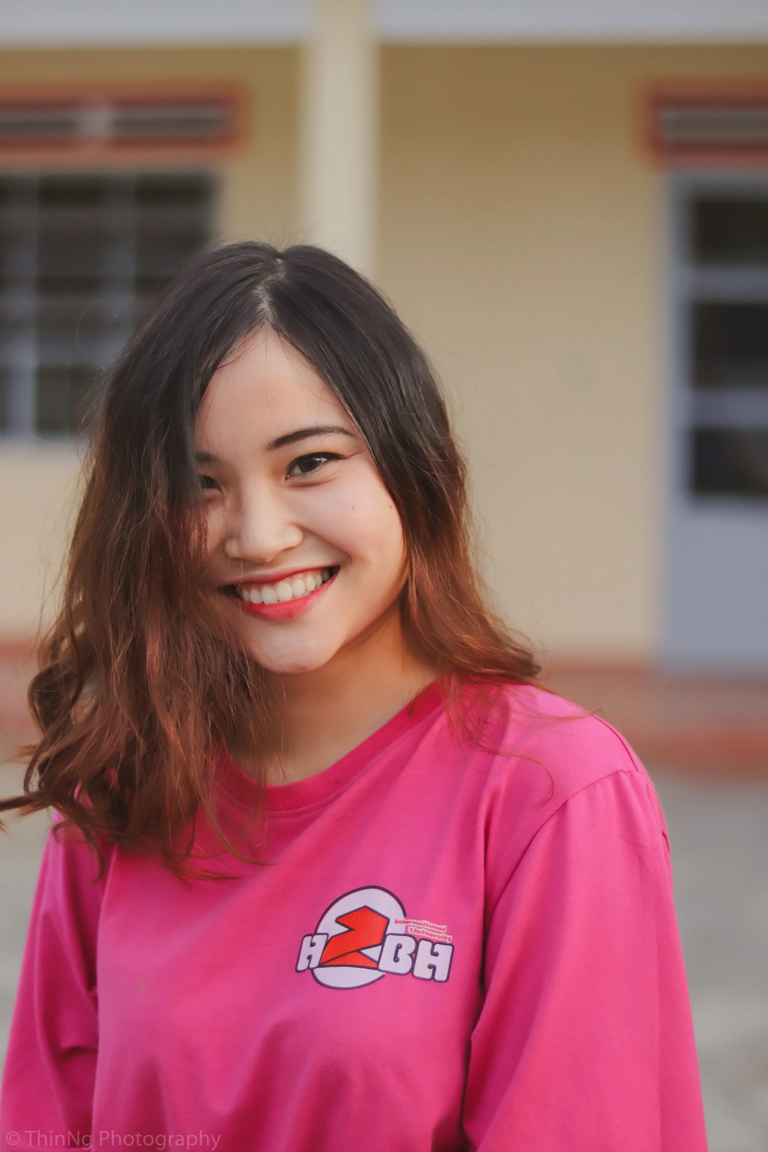 A young woman with long dark hair and a bright smile, wearing a pink shirt with a logo, standing outdoors in front of a building.