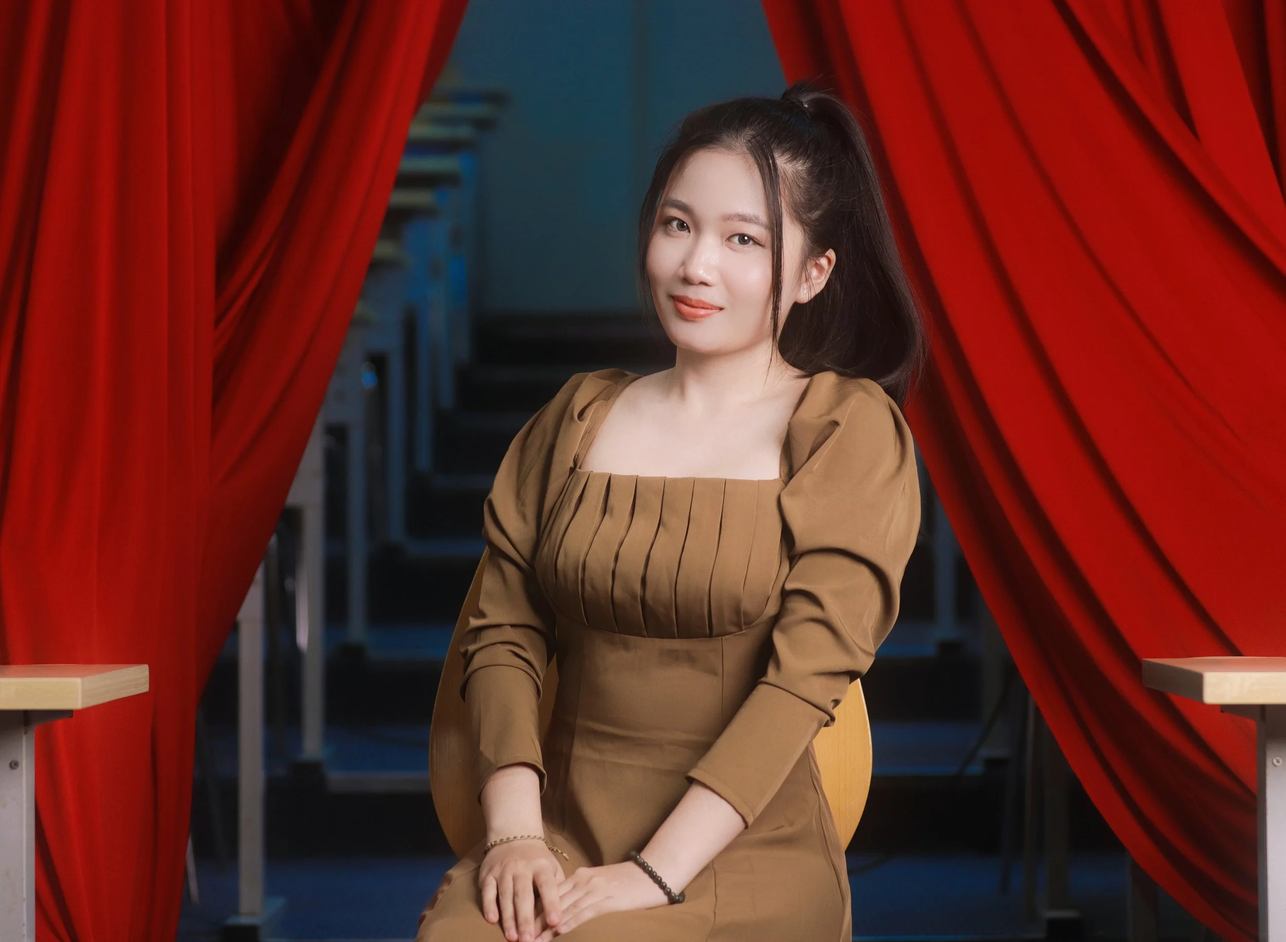 A young woman with long dark hair, sitting in front of red theater curtains, smiling at the camera.