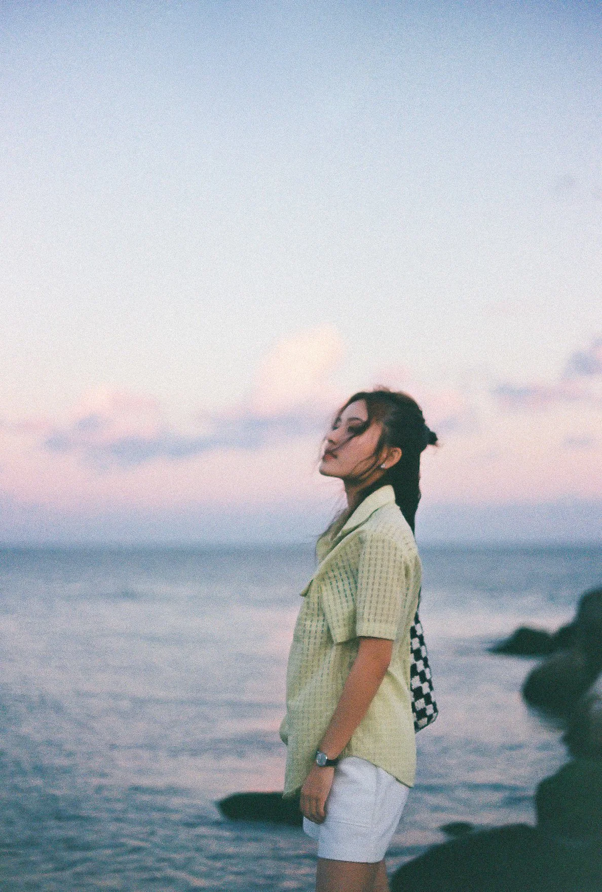 A young woman standing on rocks by the ocean during sunset, with her eyes closed and hair blowing in the wind, wearing a yellow shirt and white shorts.