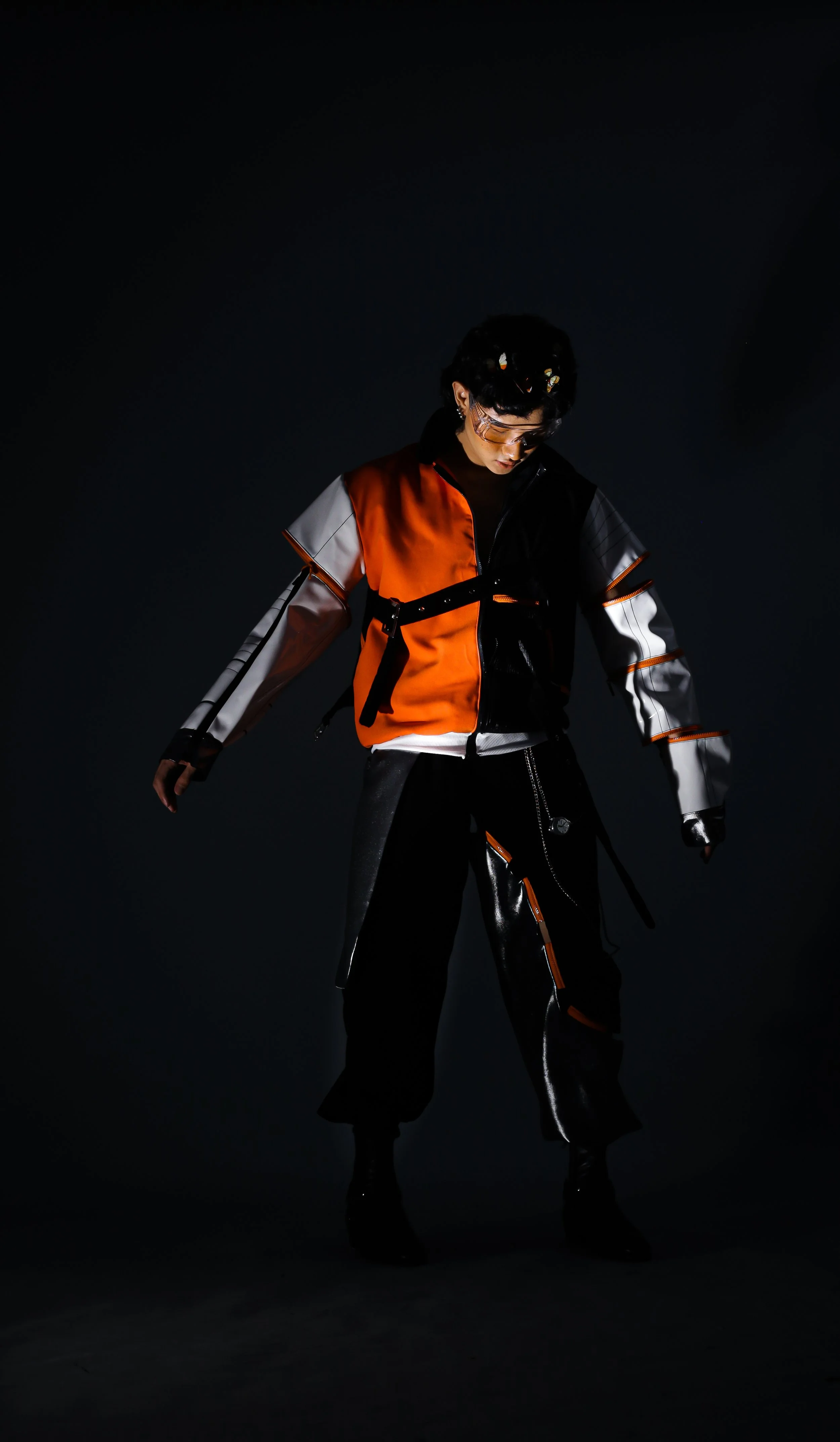 Person wearing futuristic black, orange, and white jacket and pants, standing against a dark background.