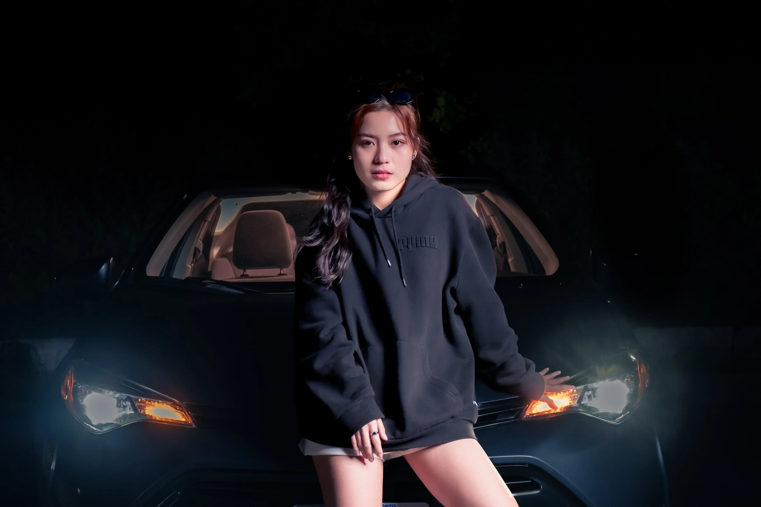 A young woman with dark wavy hair and sunglasses on her head, wearing a black hoodie, standing in front of a black car at night.