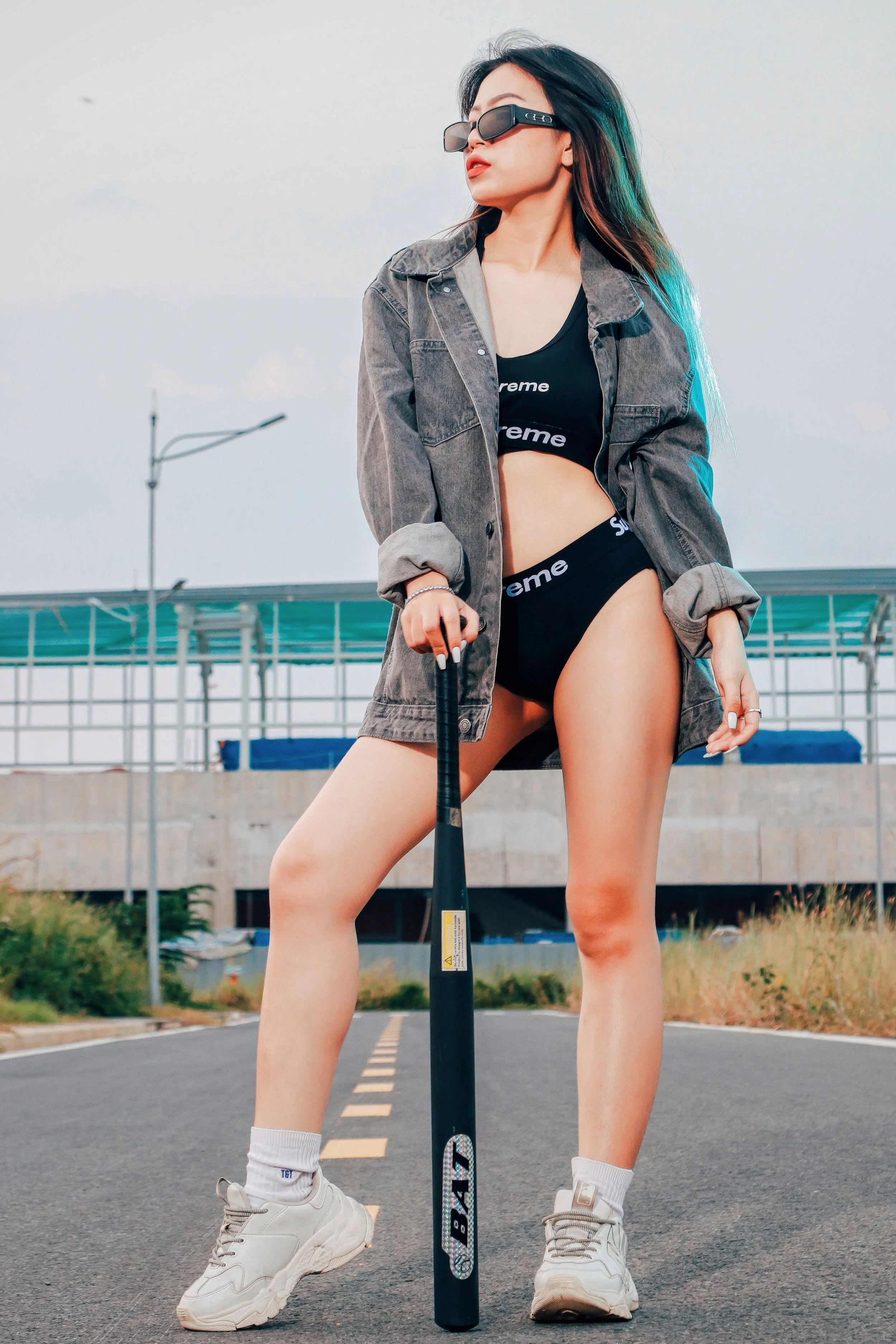 A woman standing in the middle of a road holding a baseball bat, wearing sunglasses, a black sports bra, matching underwear, a gray oversized jacket, white sneakers, and white socks with a logo, against a background of a modern bridge and a cloudy sk