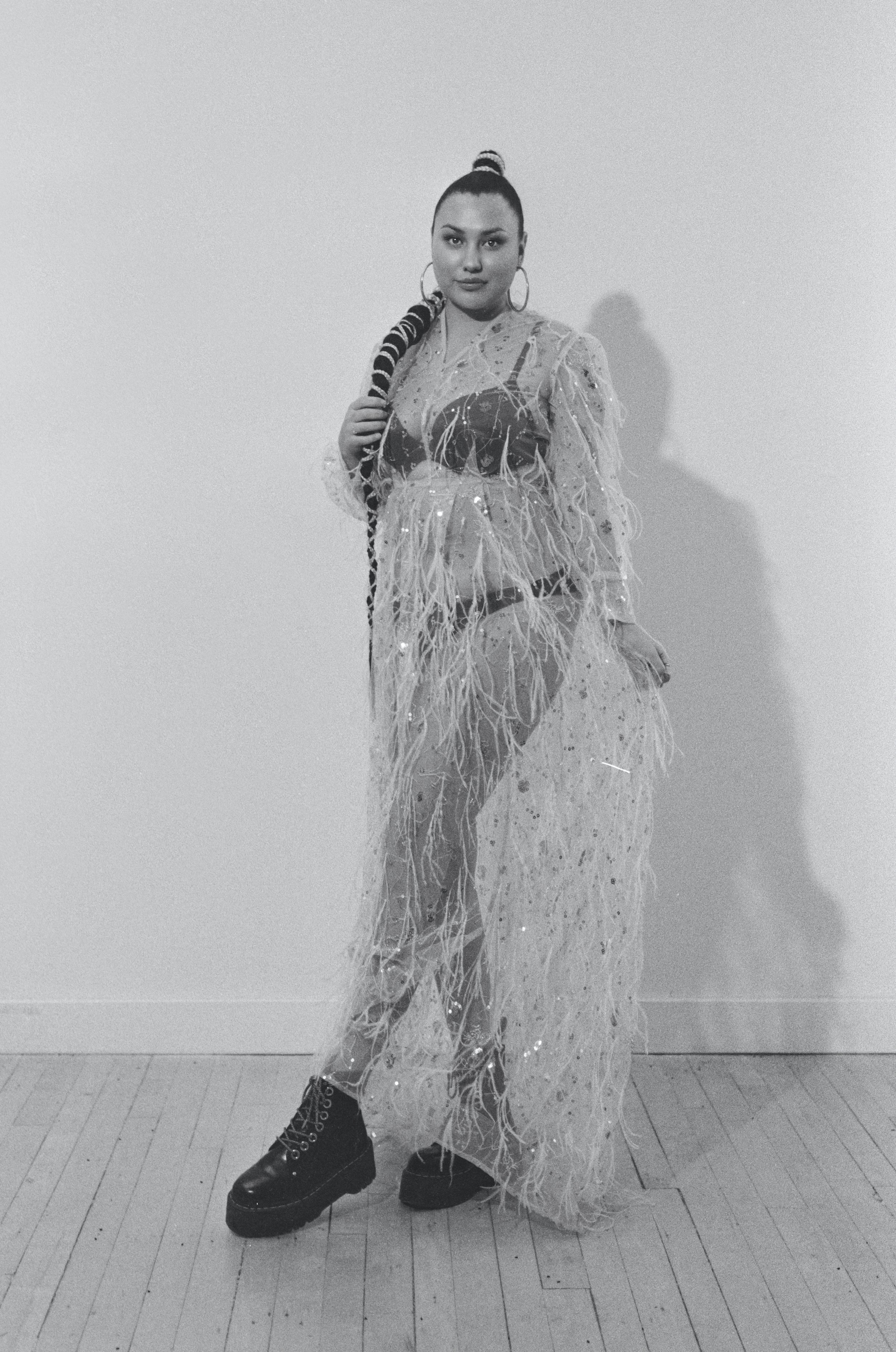 A woman in a glamorous, fringed, sheer dress with a black undergarment, chunky boots, and large hoop earrings, posing against a plain background.