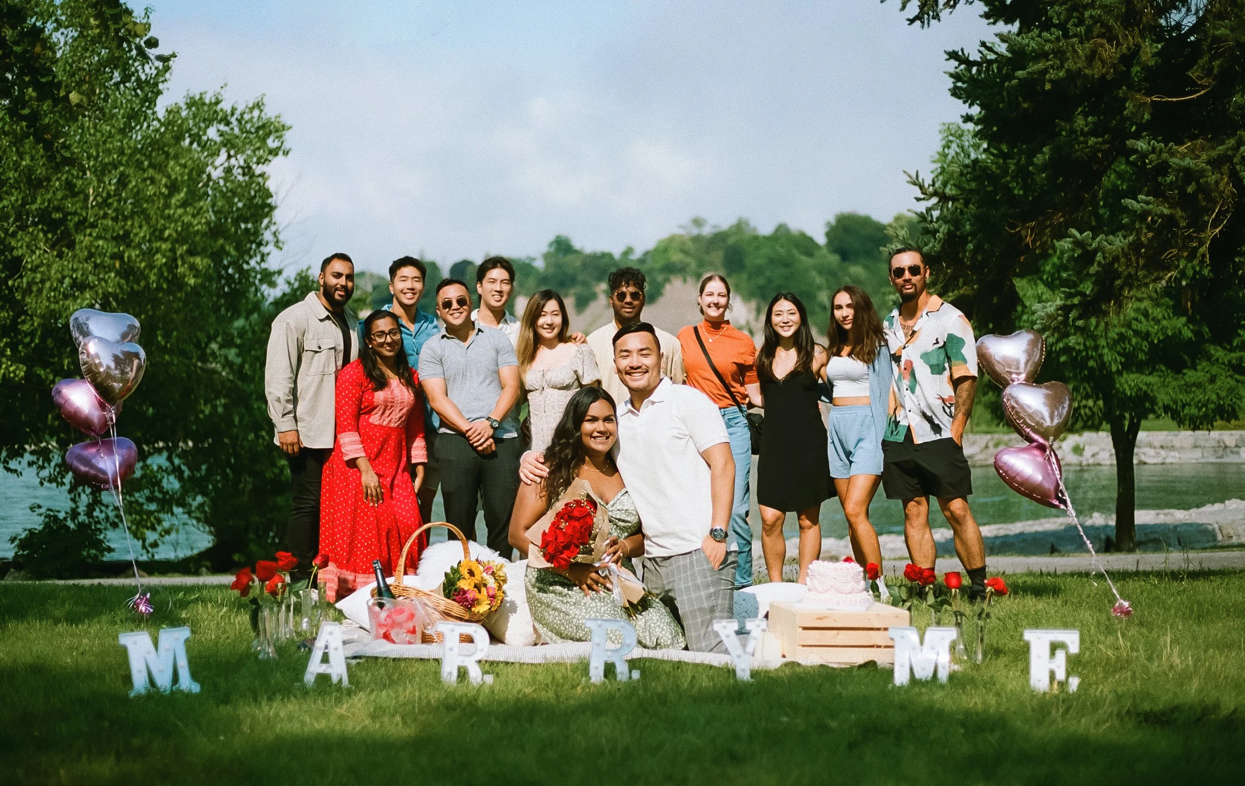 Group of people celebrating a wedding outdoors with balloons, flowers, cake, and decorations spelling out "MARRY ME" on the grass, near a river and surrounded by trees.
